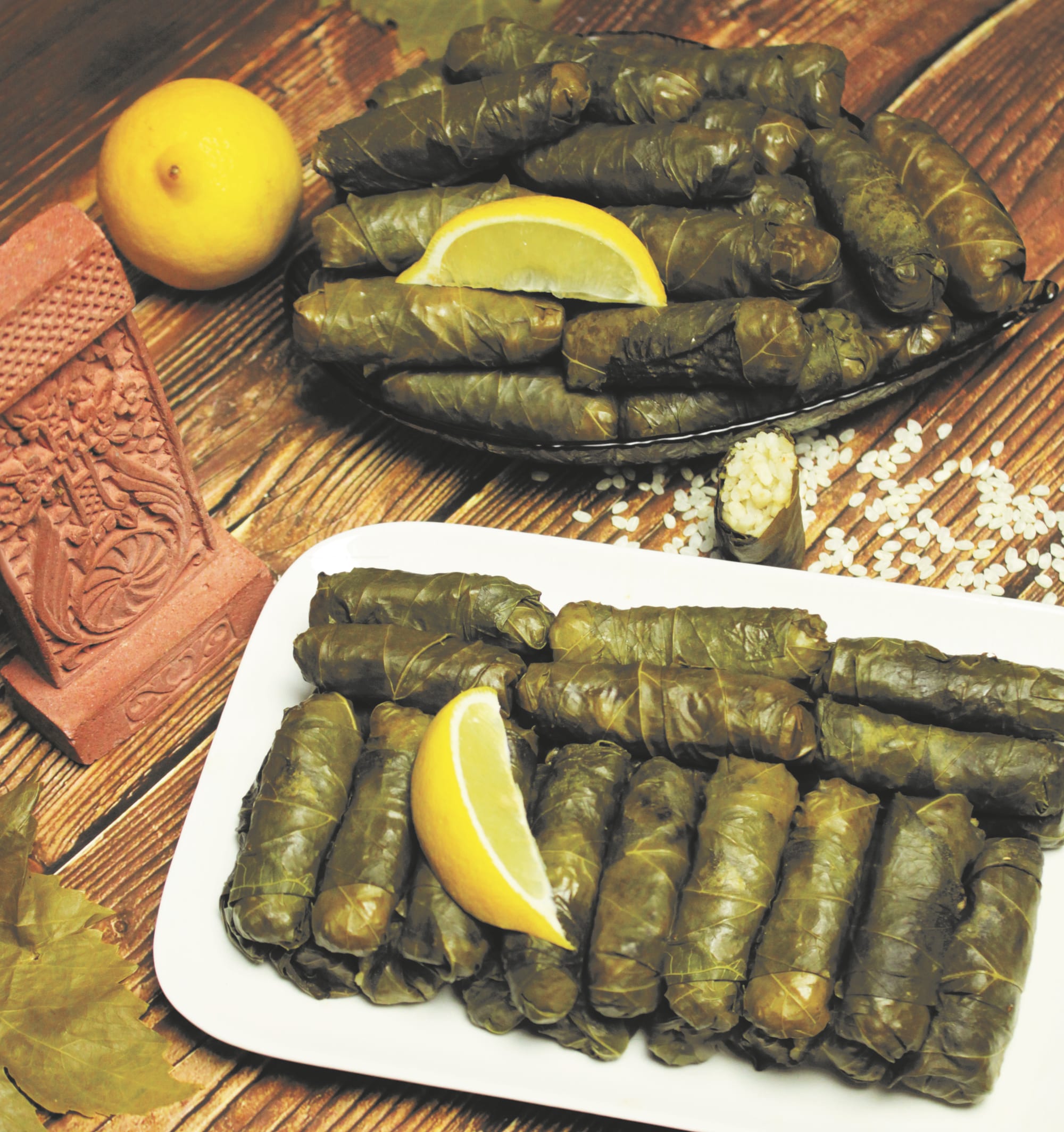 Two plates piled with small grape-leaf-wrapped rolls, garnished with lemon wedges, one roll cut open to show a rice filling