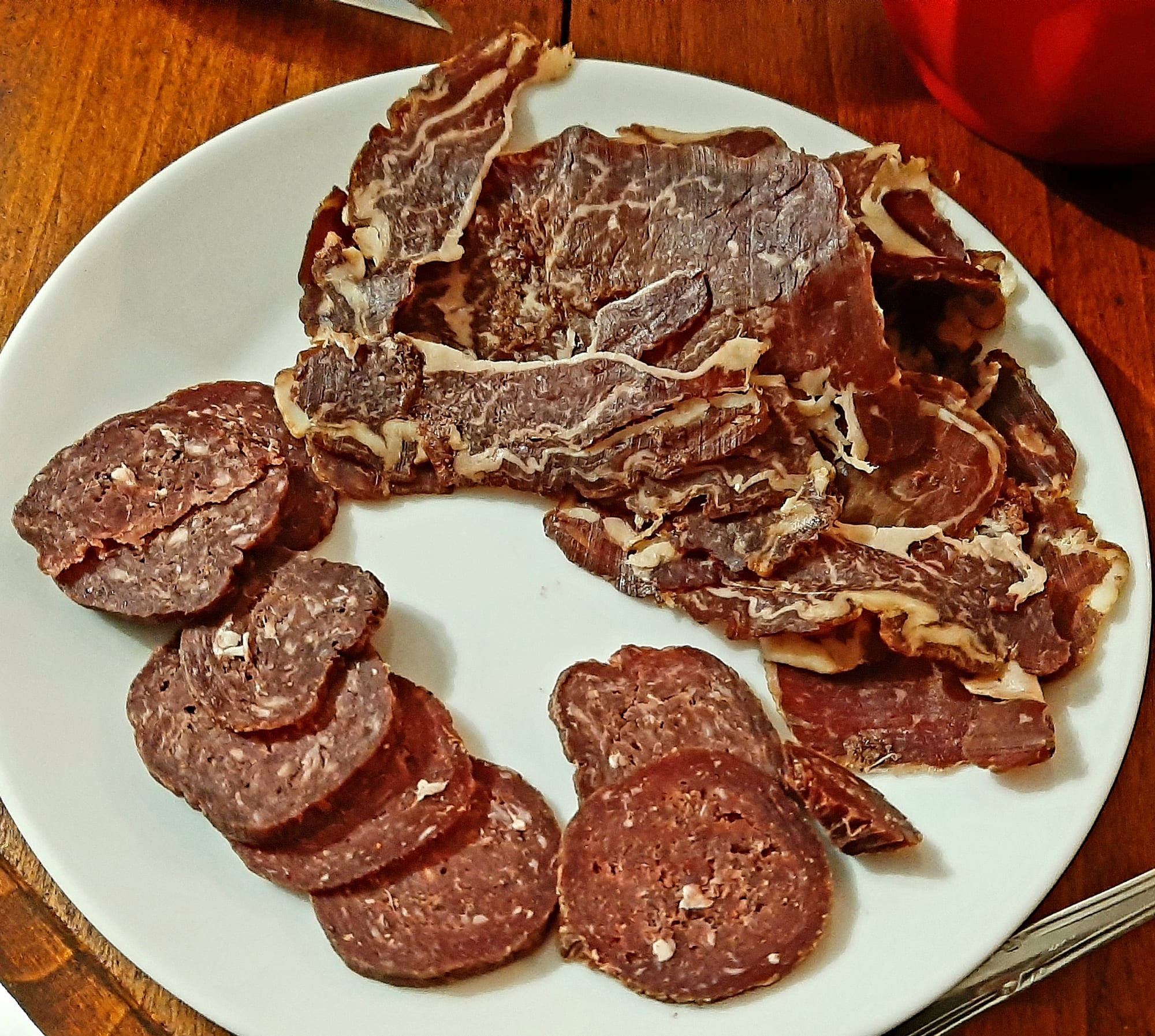 A white plate holding dark, spice-crusted dried beef slices and dark round slices of cured sausage