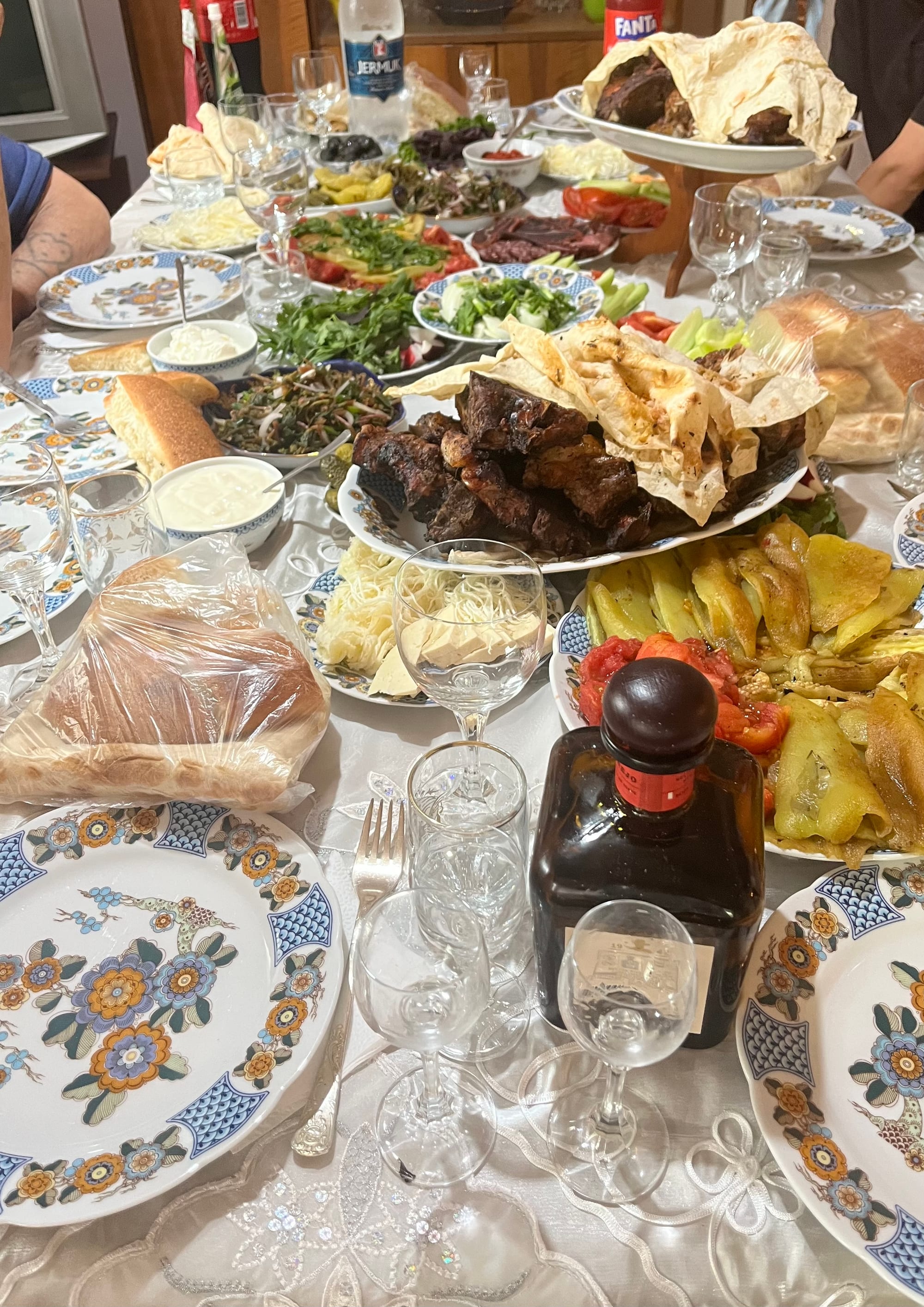 A table crowded with plates of grilled meat, lavash bread, fresh herbs, sliced tomatoes, pickled peppers and wine glasses