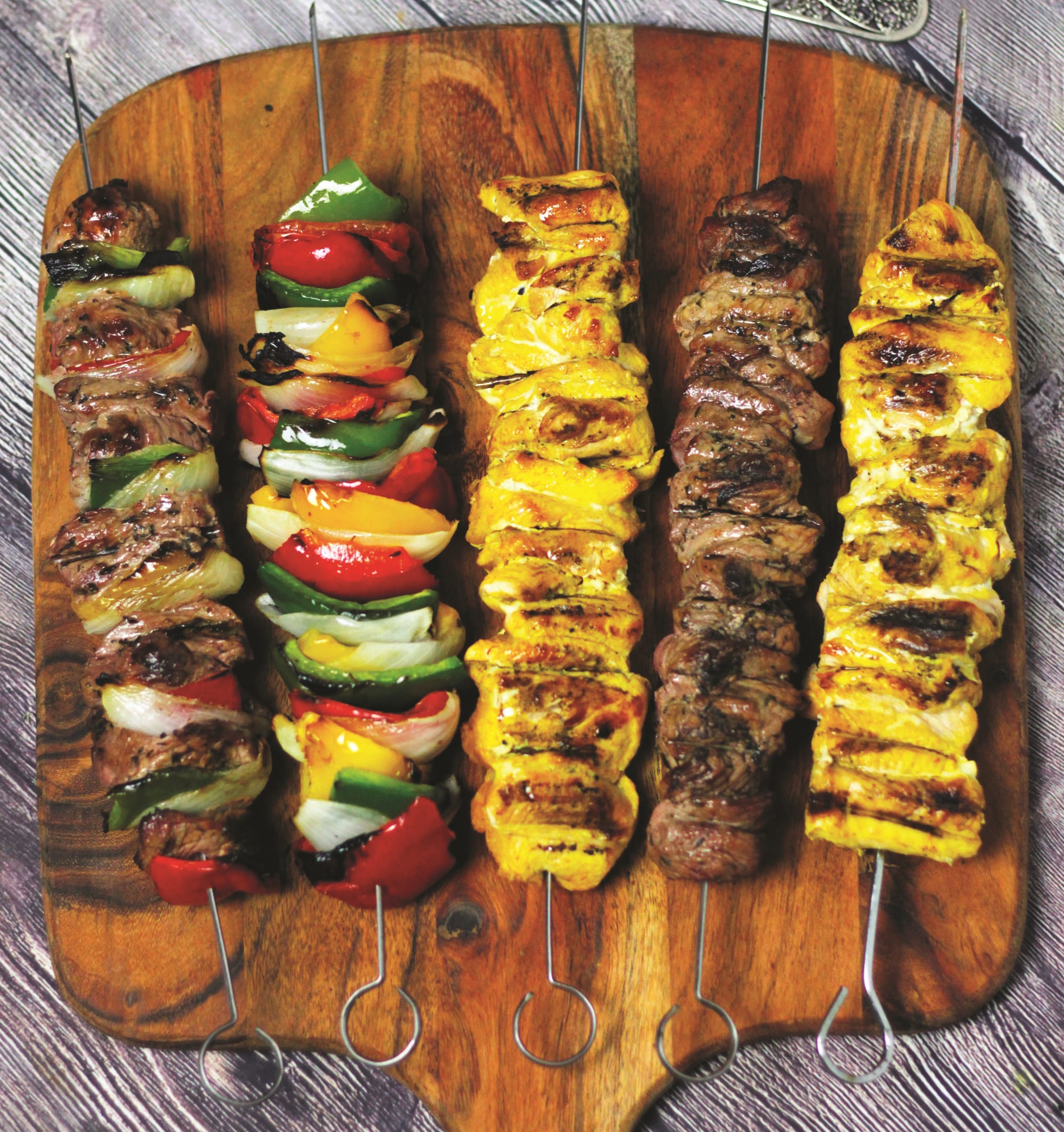 Five long metal skewers of grilled meat and vegetables laid across a wooden board