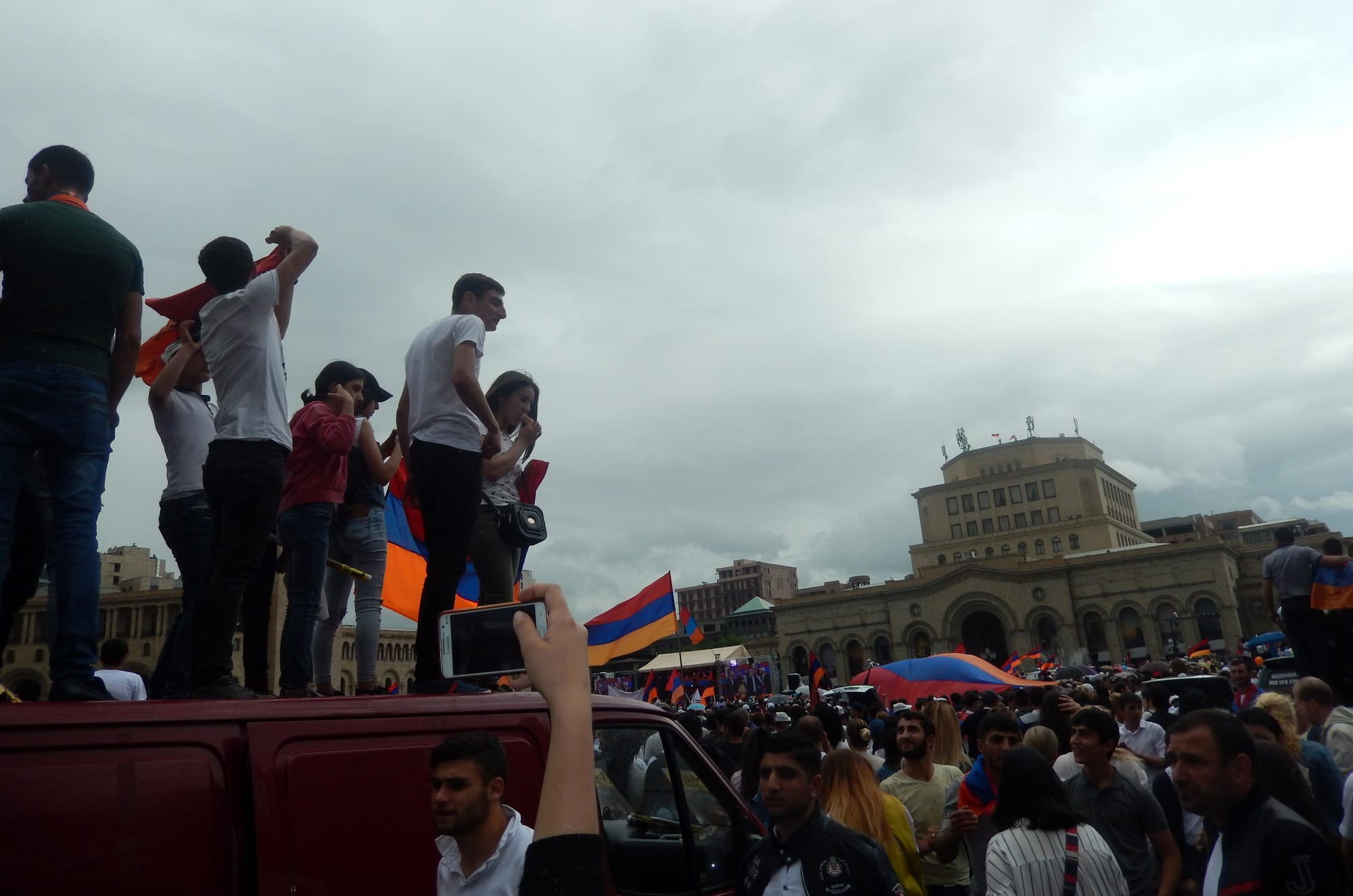 A packed city square at dusk full of people, many holding up phones and Armenian flags