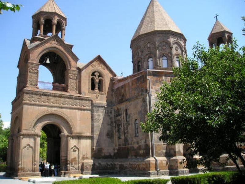 A compact stone cathedral with a conical central dome and a tall bell tower, among trees