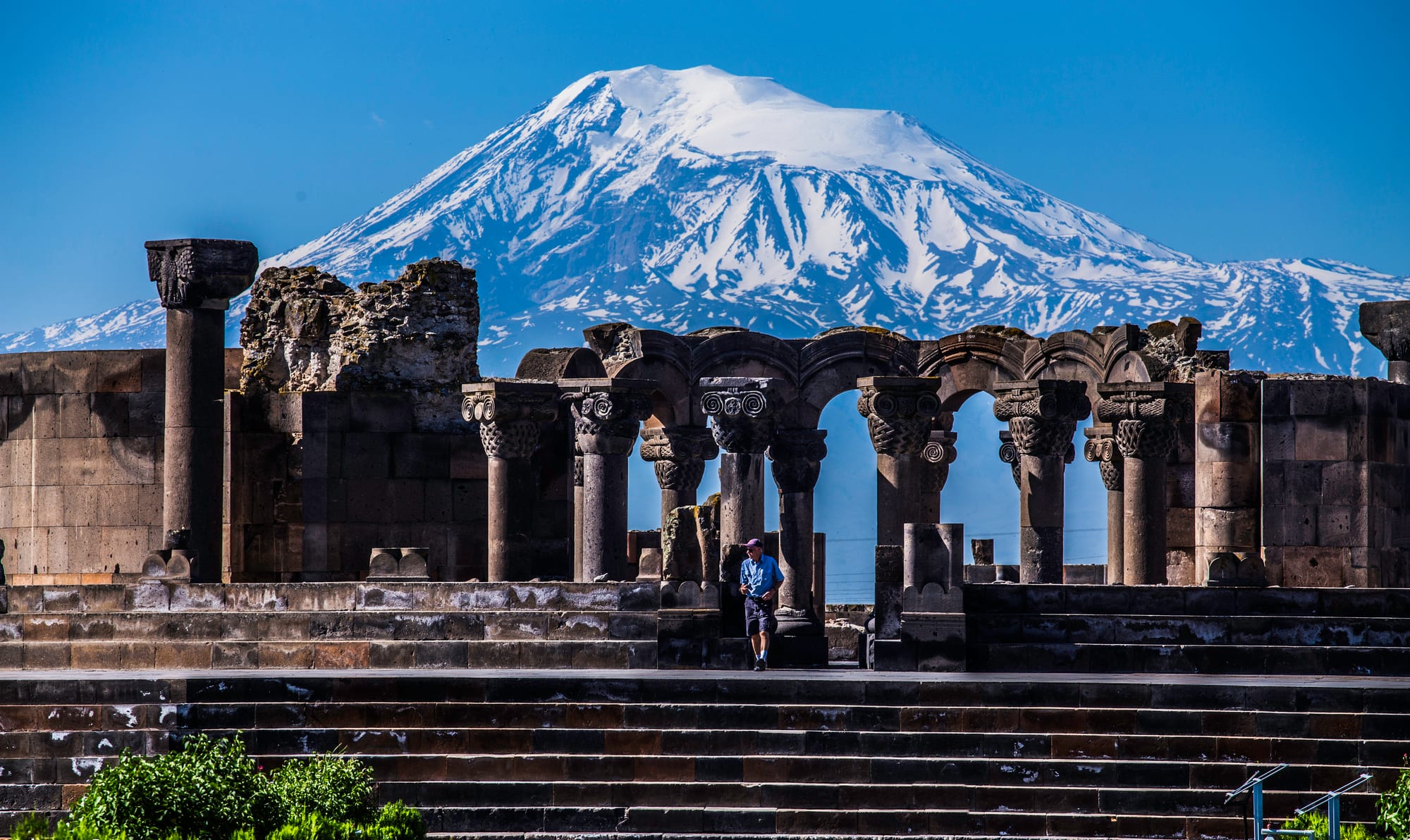 A ruined circle of dark stone columns and arches on a stepped platform, with the snow-covered cone of Mount Ararat filling the sky behind