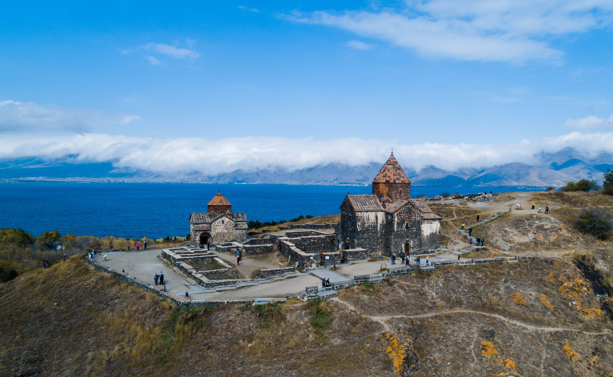 Two small stone churches with conical domes on a bare headland above a wide blue lake, with mountains under cloud on the far shore