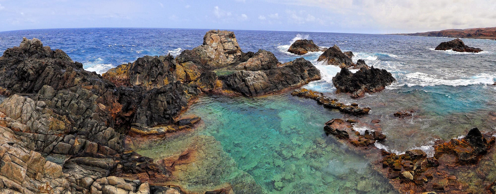 A circular pool of clear water enclosed by a ring of black volcanic rocks, waves breaking outside