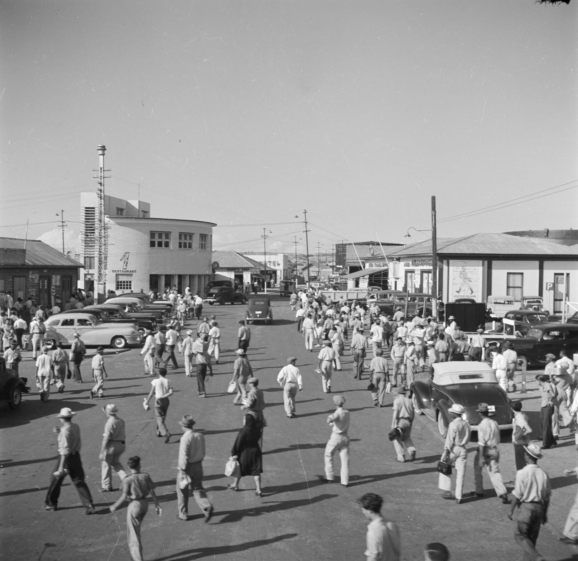 A 1940s black-and-white photograph of a large crowd of workers streaming out of a refinery gate on bicycles and on foot