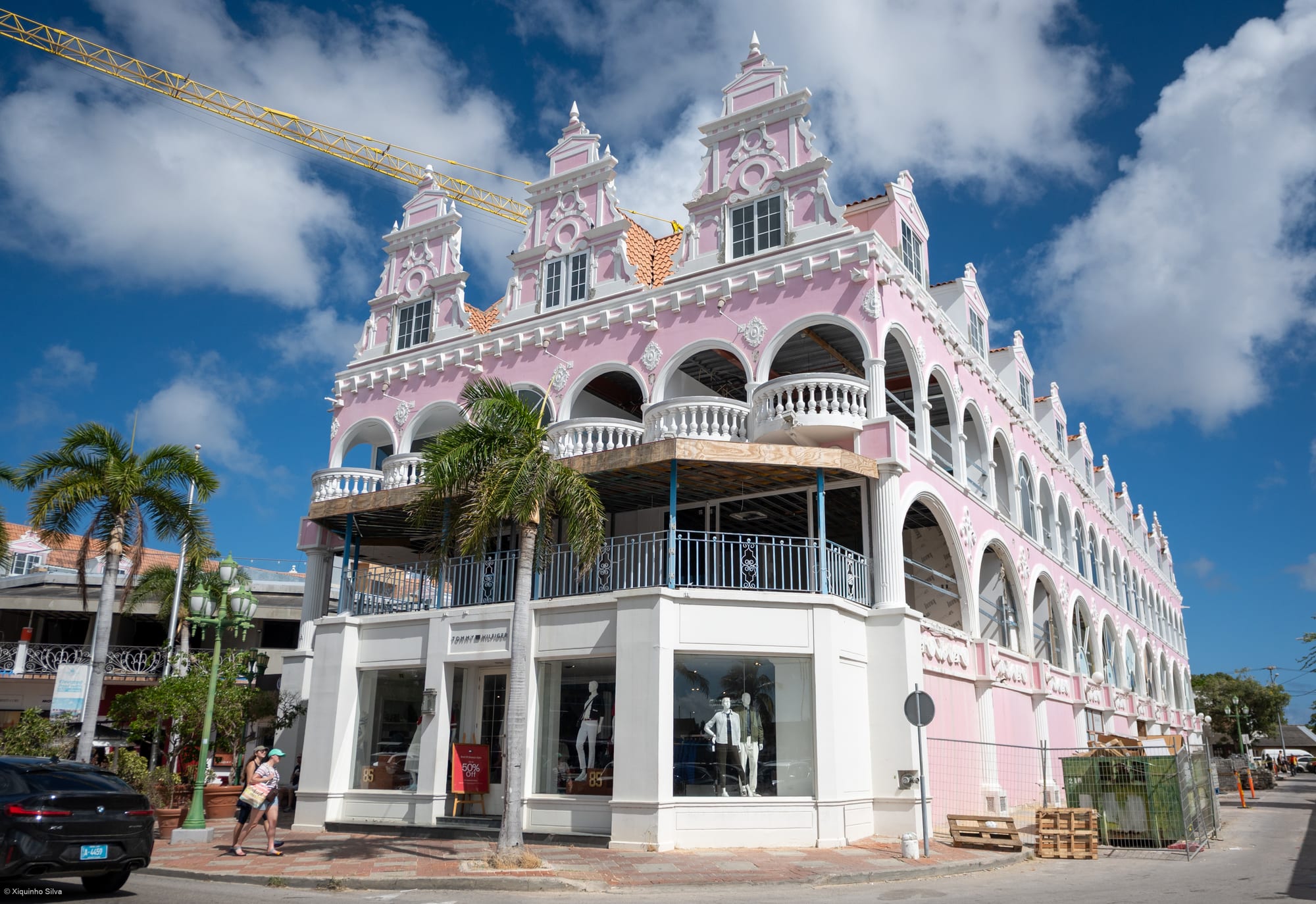 A pink and white Dutch colonial-style building with ornate gables and arcades in Oranjestad, palm trees in front and pedestrians walking past the shopfronts