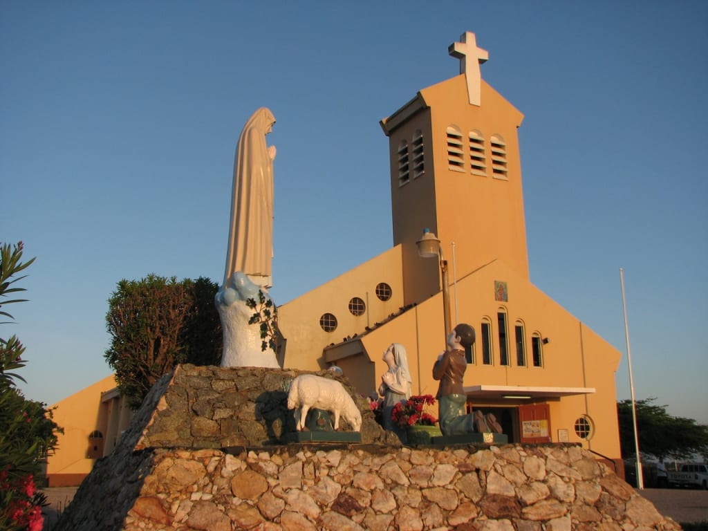 An orange stone parish church with a bell tower and cross, statues of the Virgin Mary and kneeling figures in front, lit by evening light