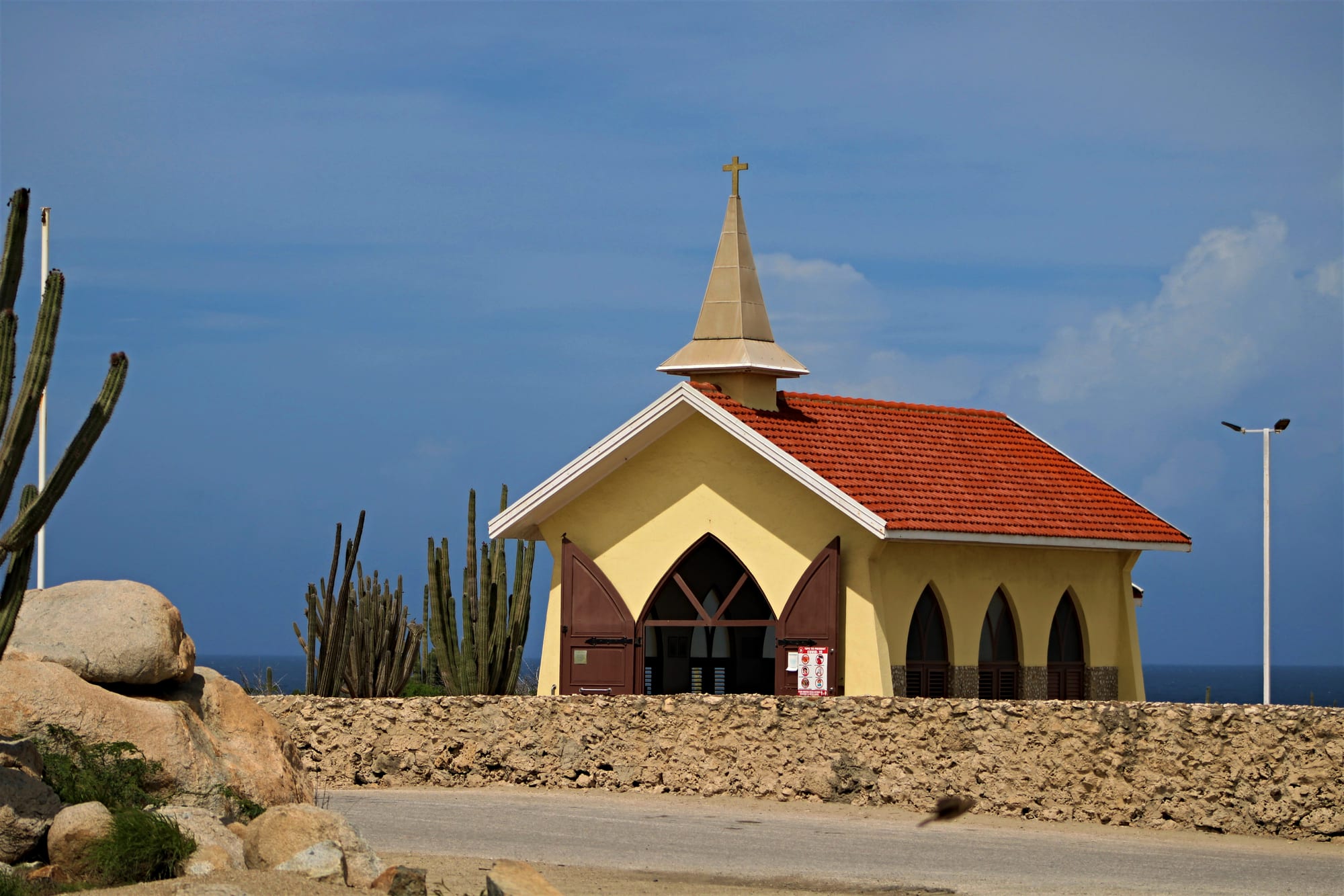 A small yellow chapel with a red tiled roof, a pointed spire topped by a cross and brown pointed-arch doors, behind a low stone wall with tall cacti and boulders nearby and the sea on the horizon