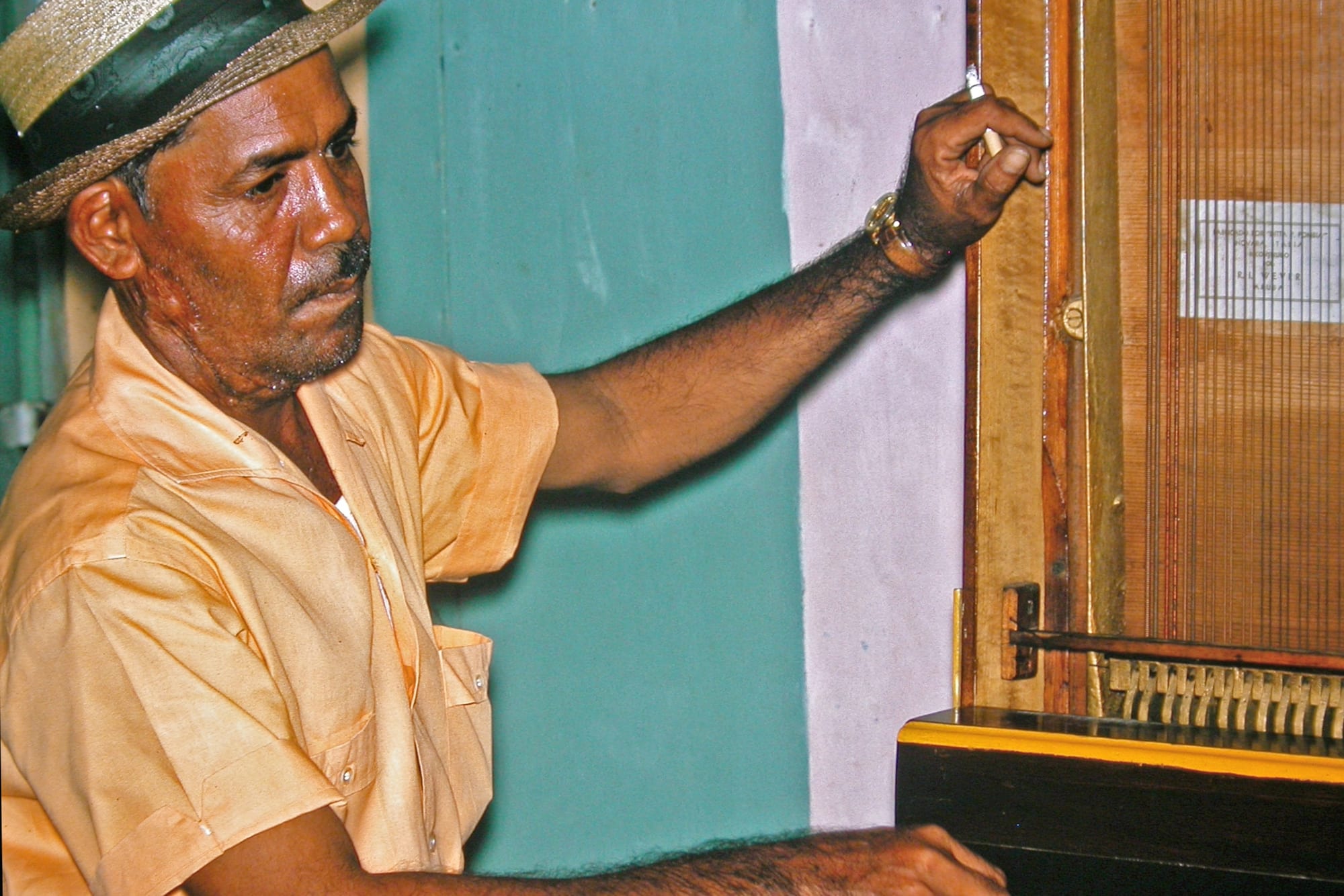 A man in a straw hat and an orange shirt stands beside a tall wooden barrel organ, one hand resting on its glass-fronted case, against a green and white wall