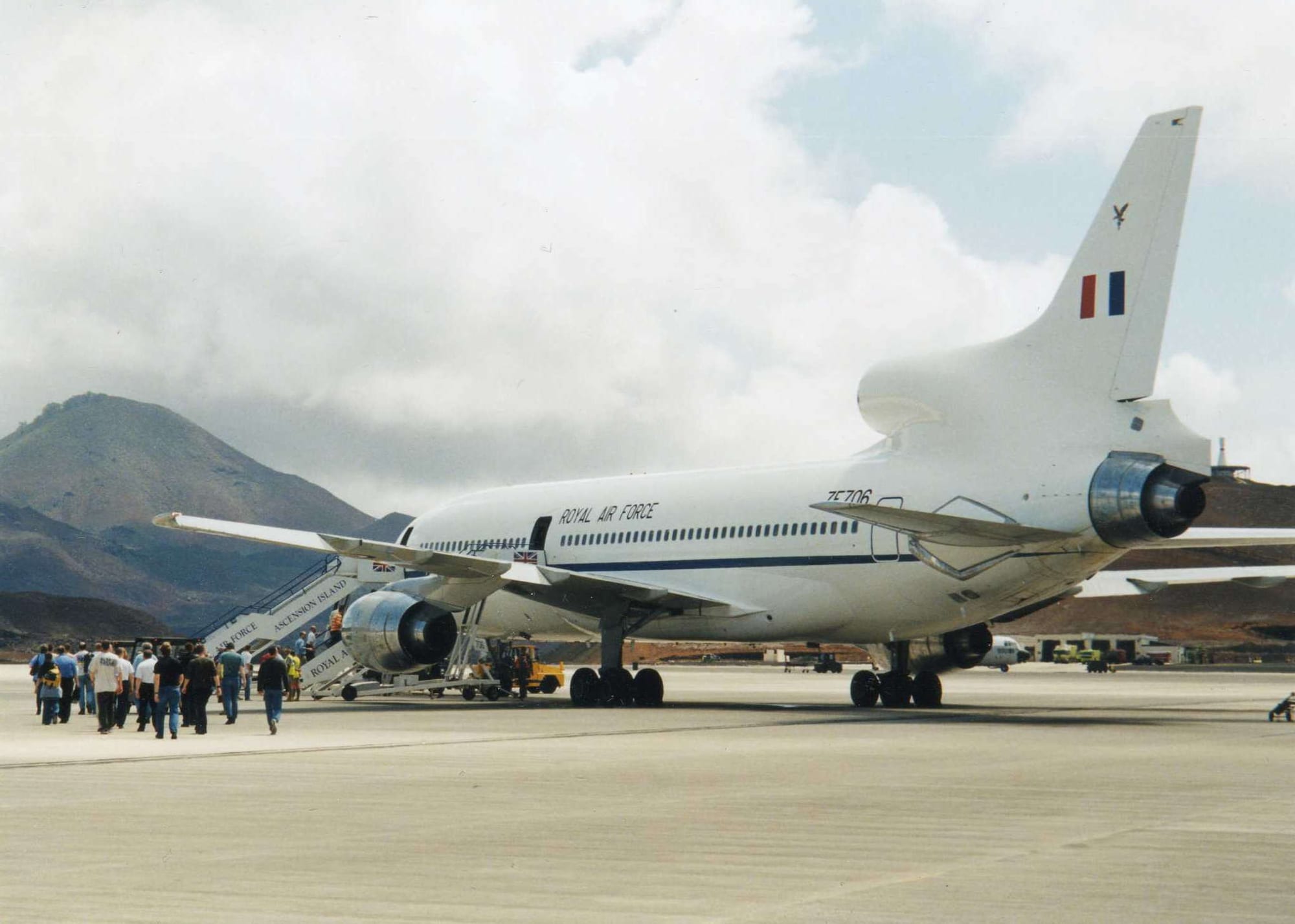 A large RAF transport jet parked on an apron under a bright sky with a volcanic hill behind