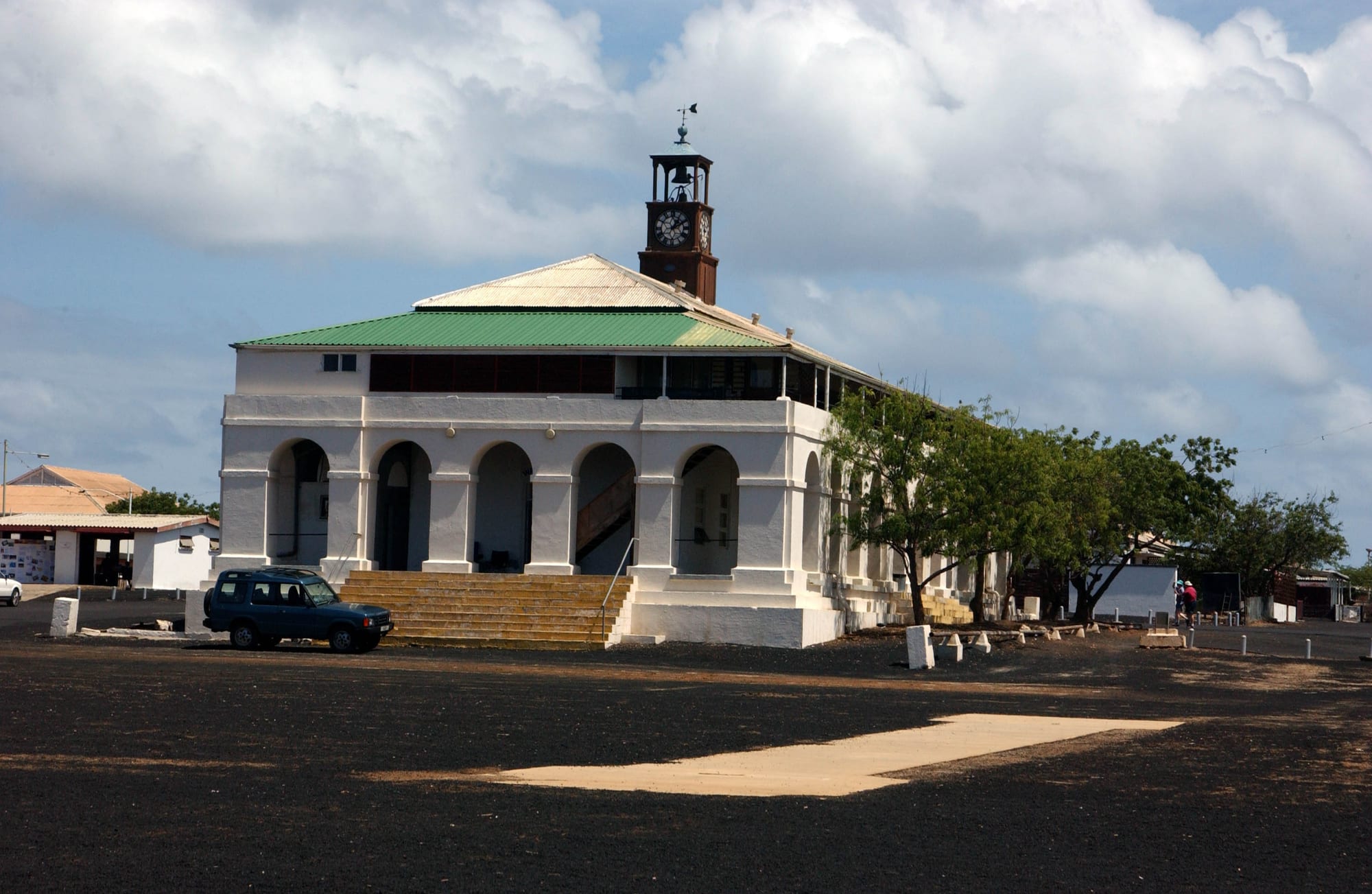 A white colonial-style civic building with a green roof and a clock tower, a parked Land Rover outside on bare black volcanic ground