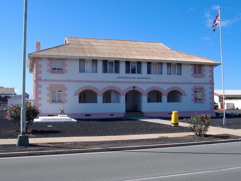 A two-storey pink and white colonial-style government building with arched arcades and a British flag flying beside it, on bare black volcanic ground