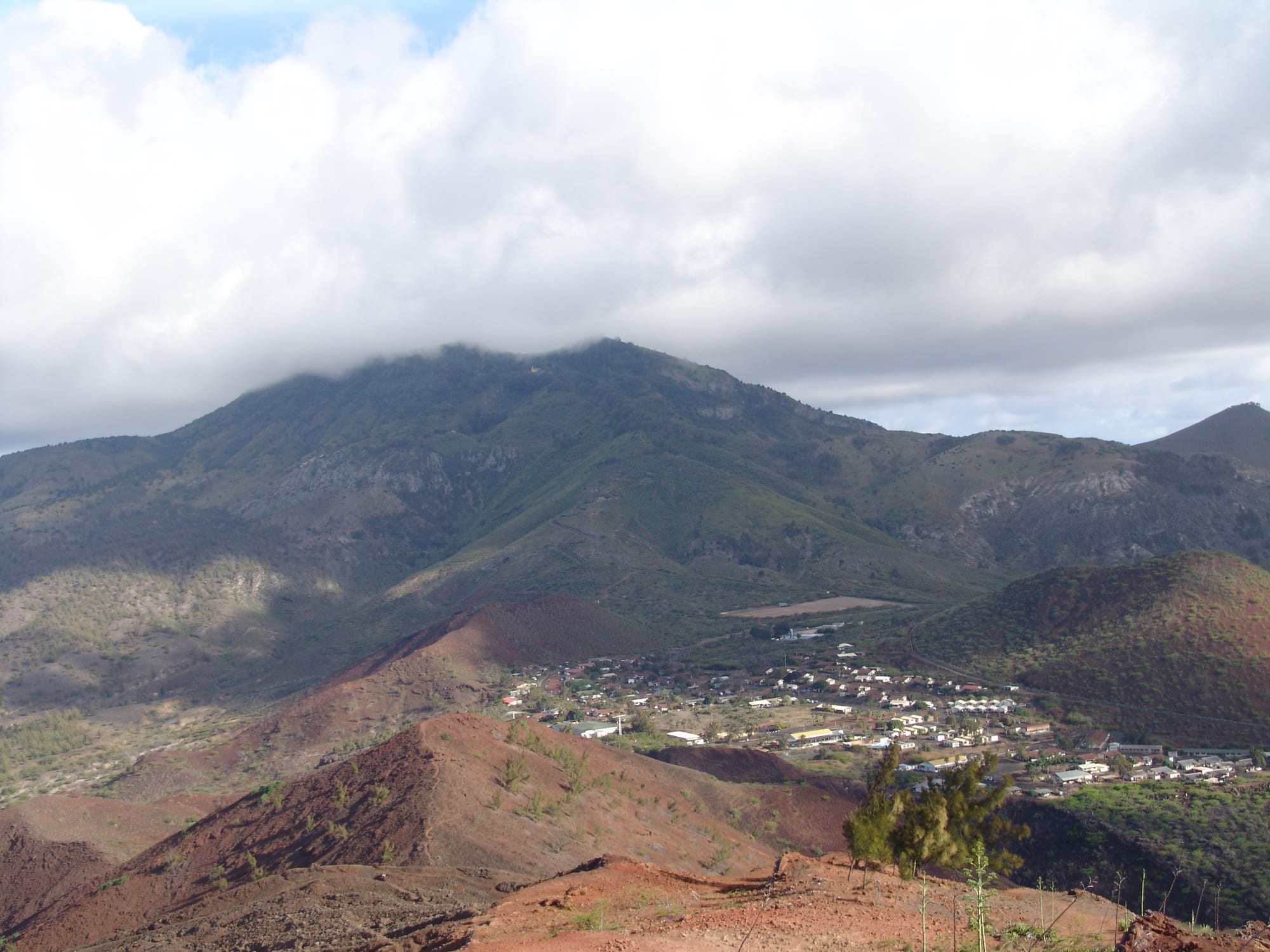 A small cluster of white-roofed houses making up Two Boats village, seen from above on a red volcanic slope, with the cloud-wrapped green summit of Green Mountain behind