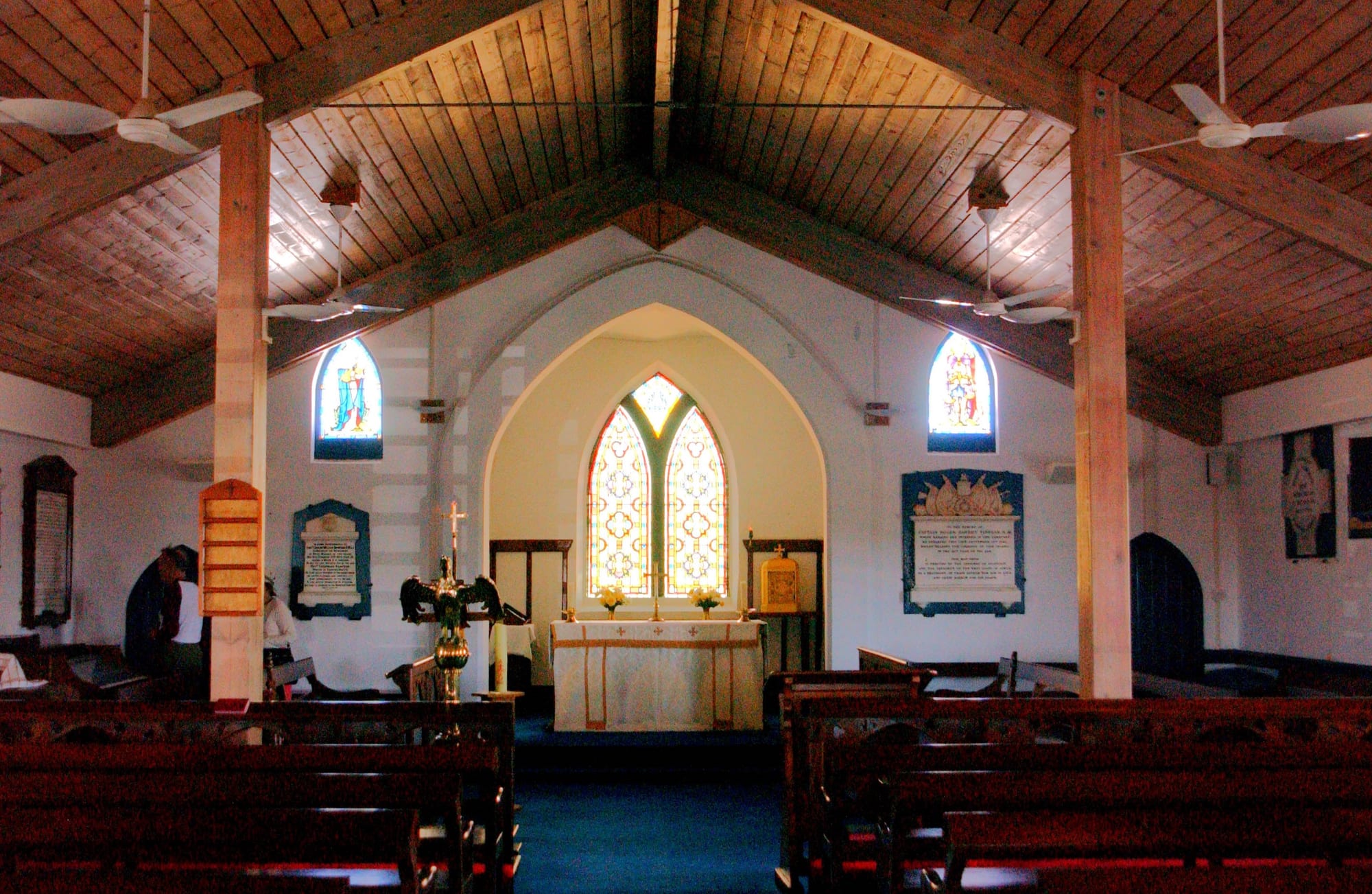 The interior of a small church with a pitched timber ceiling, ceiling fans, wooden pews, memorial plaques on white walls and stained glass windows above a simple altar