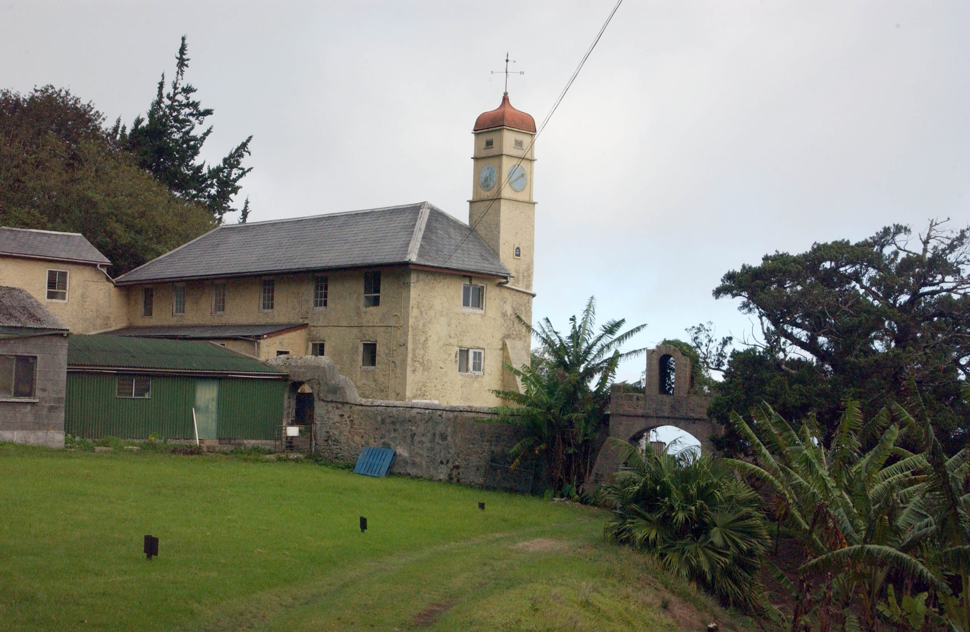 A pale two-storey building with a slate roof and a tall clock tower topped by a small dome, beside a stone wall with an arched gateway, a lawn in front and trees and banana plants around it