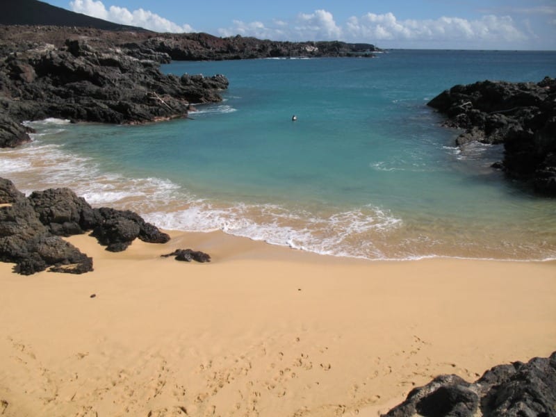 A small sandy cove between black lava headlands with clear blue water