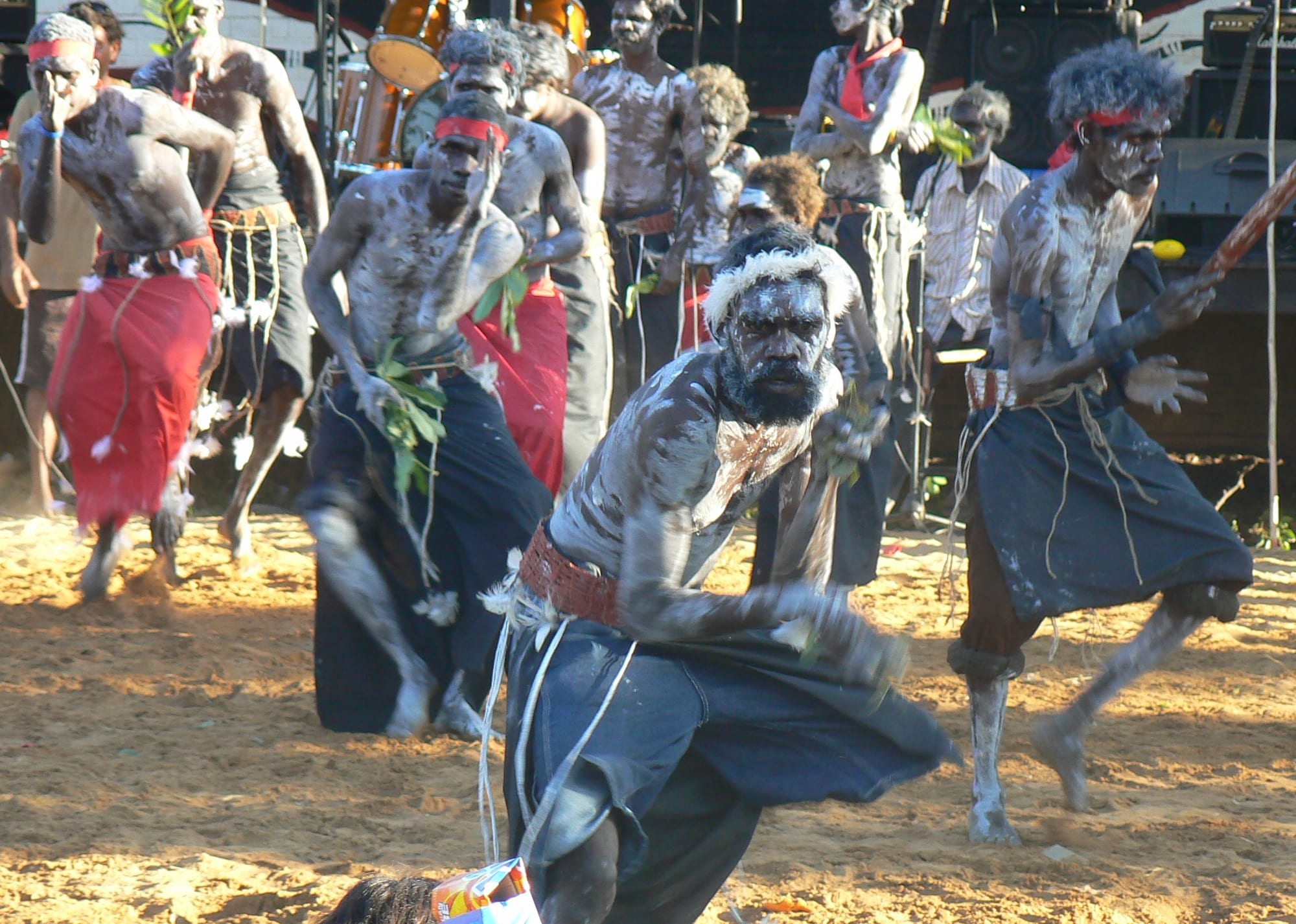 A line of Aboriginal men with white body paint, red headbands and cloth skirts dance on red sand, one in the foreground crouching low with a stick in his hand