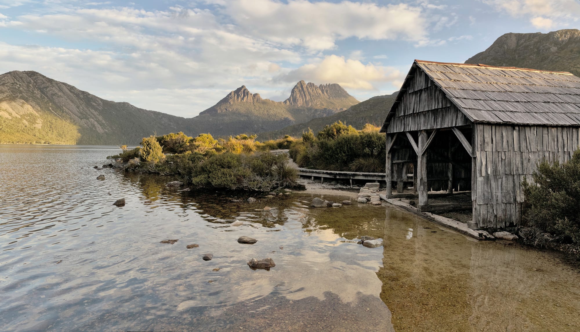 A jagged mountain reflected in a still lake, an old wooden boat shed on the near shore