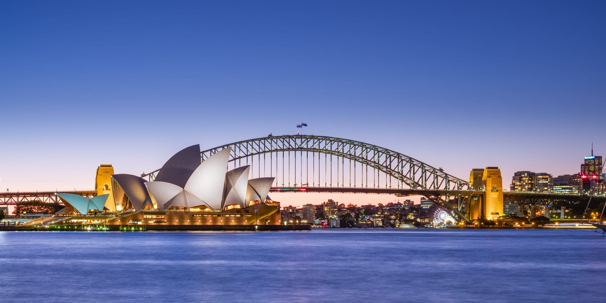 The white shell roofs of the Opera House with the steel arch of the Harbour Bridge behind at dusk
