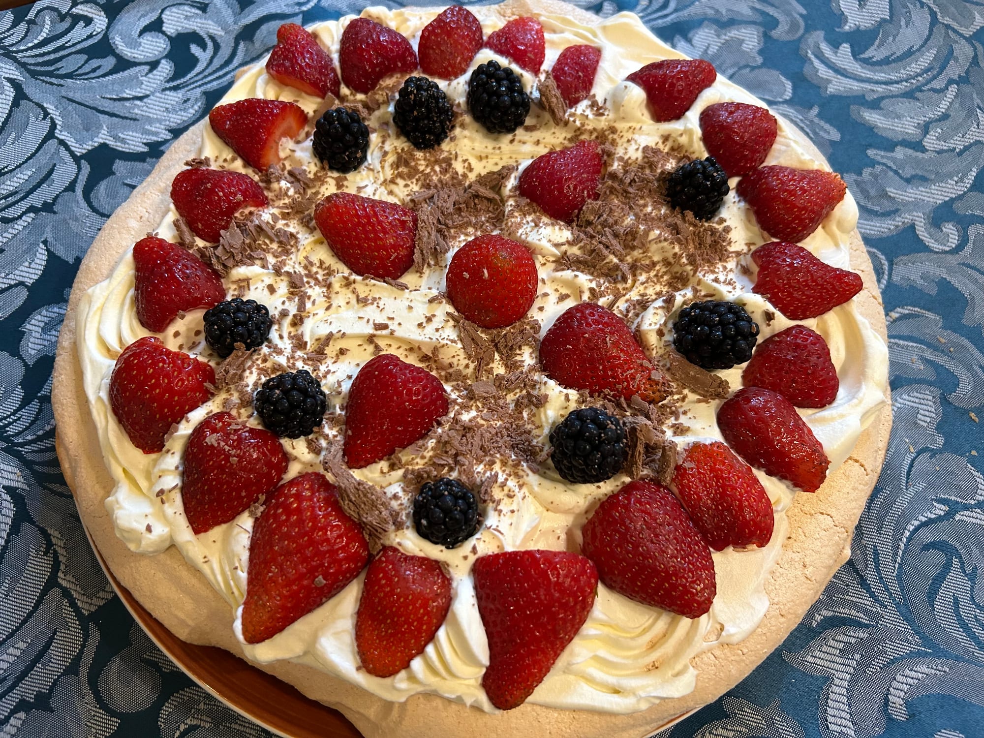 A round meringue dessert topped with whipped cream, strawberries, blackberries and shaved chocolate