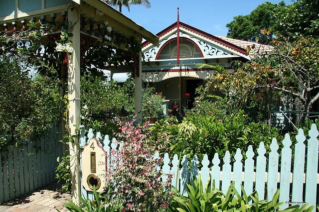 A timber cottage with a decorative gable and verandah, behind a white picket fence and a garden of flowering plants