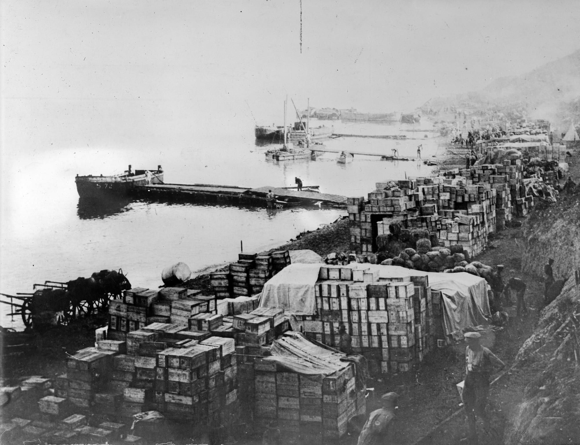 Crates and barrels of supplies stacked along a narrow beach below steep cliffs, with boats offshore