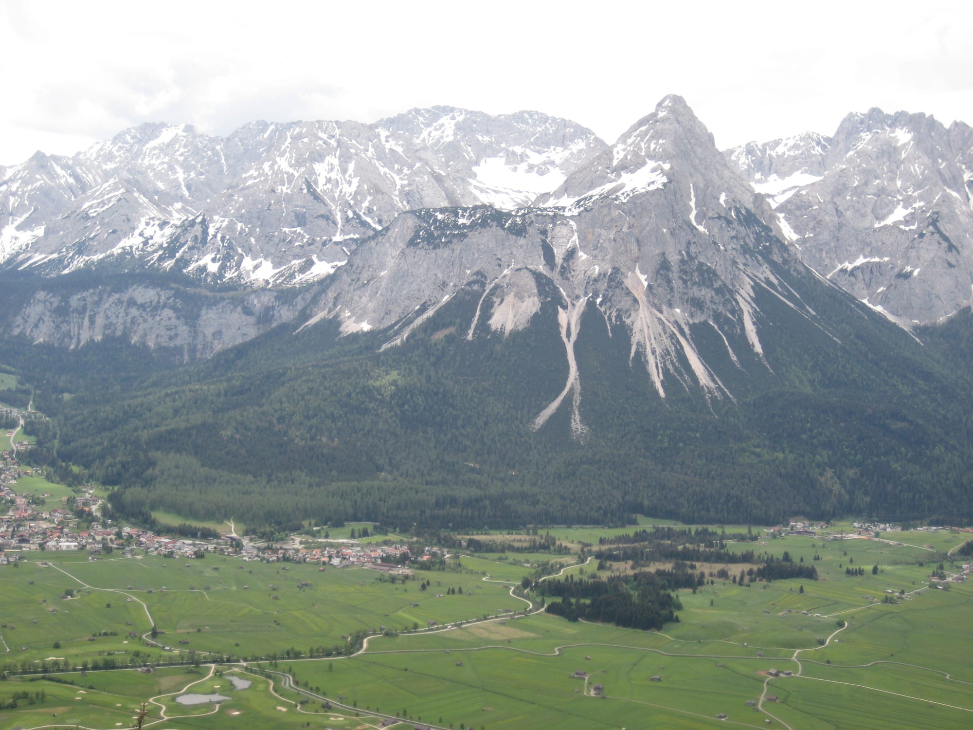 A wide alpine valley with a small village, green fields and forest below a range of snow-streaked mountains