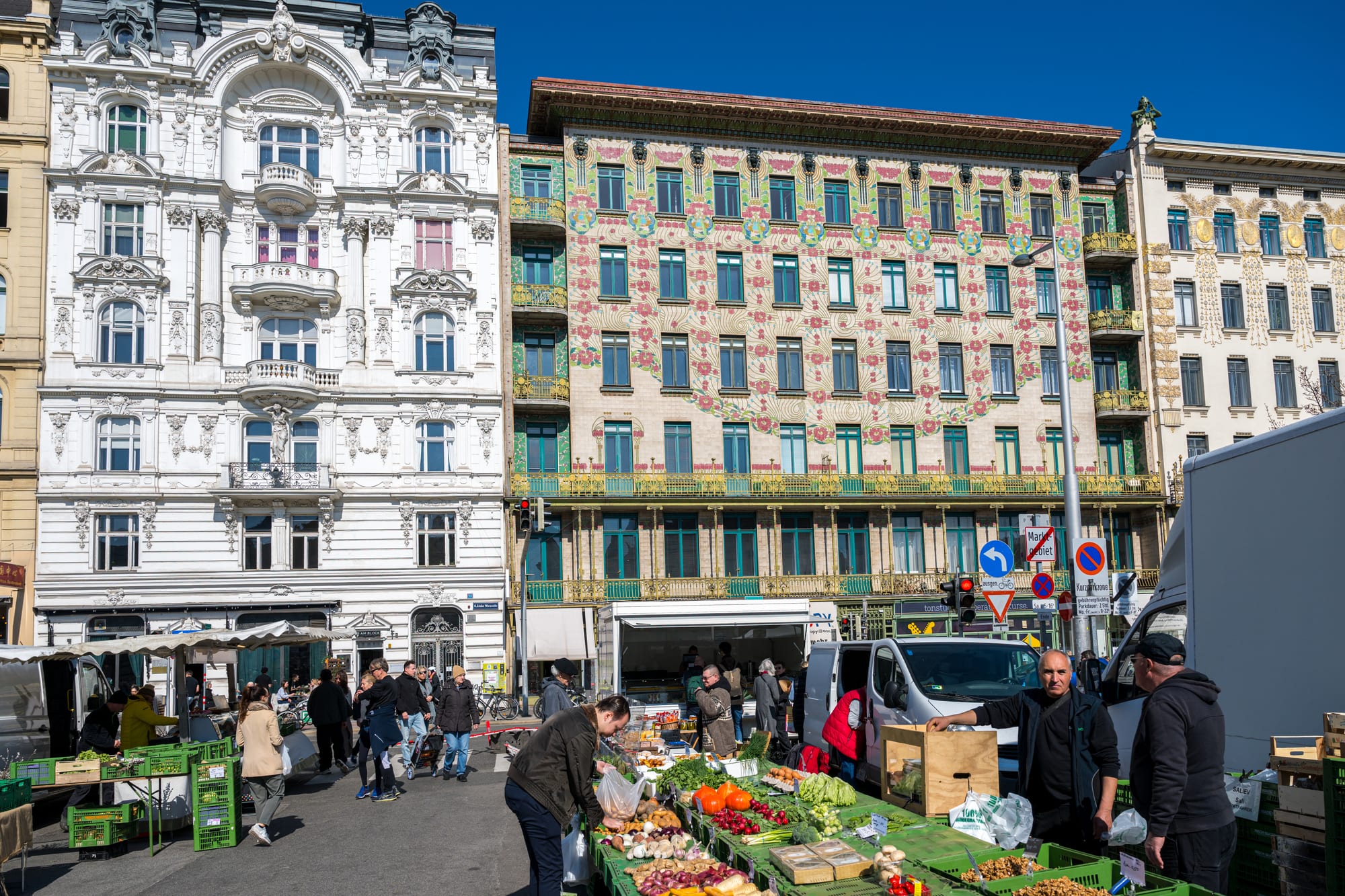 A busy outdoor market street lined with ornate historic buildings, with vendors and shoppers browsing stalls of fresh vegetables