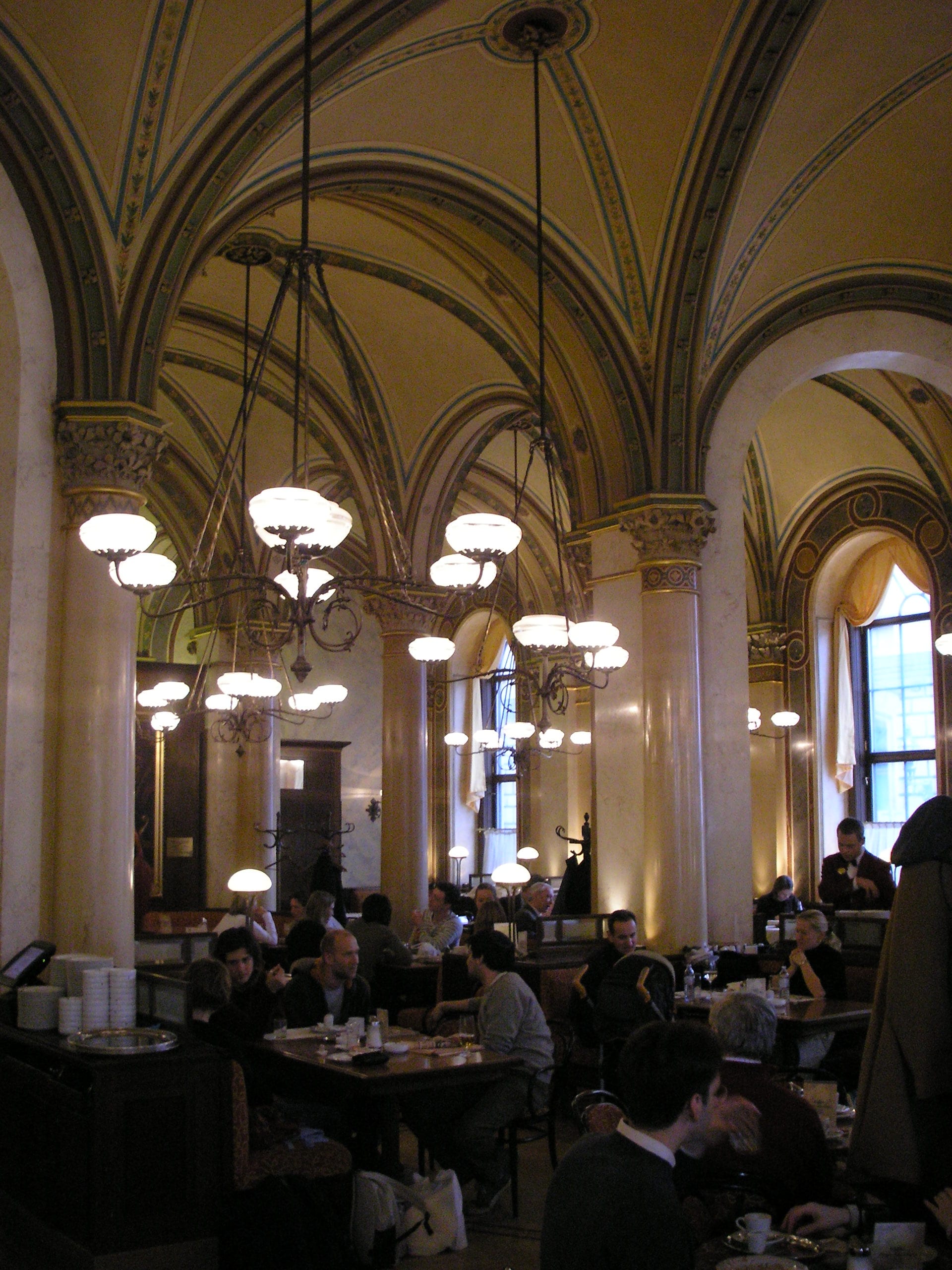 A grand vaulted coffeehouse interior with ornate columns, hanging chandeliers and groups of people seated at tables