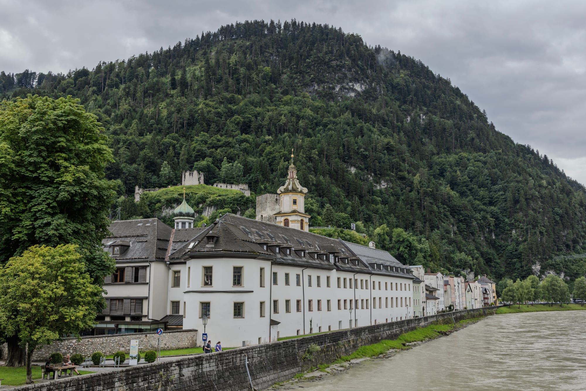 A row of pastel and white townhouses with a church tower along the Inn river, beneath a steep forested hill topped with castle ruins