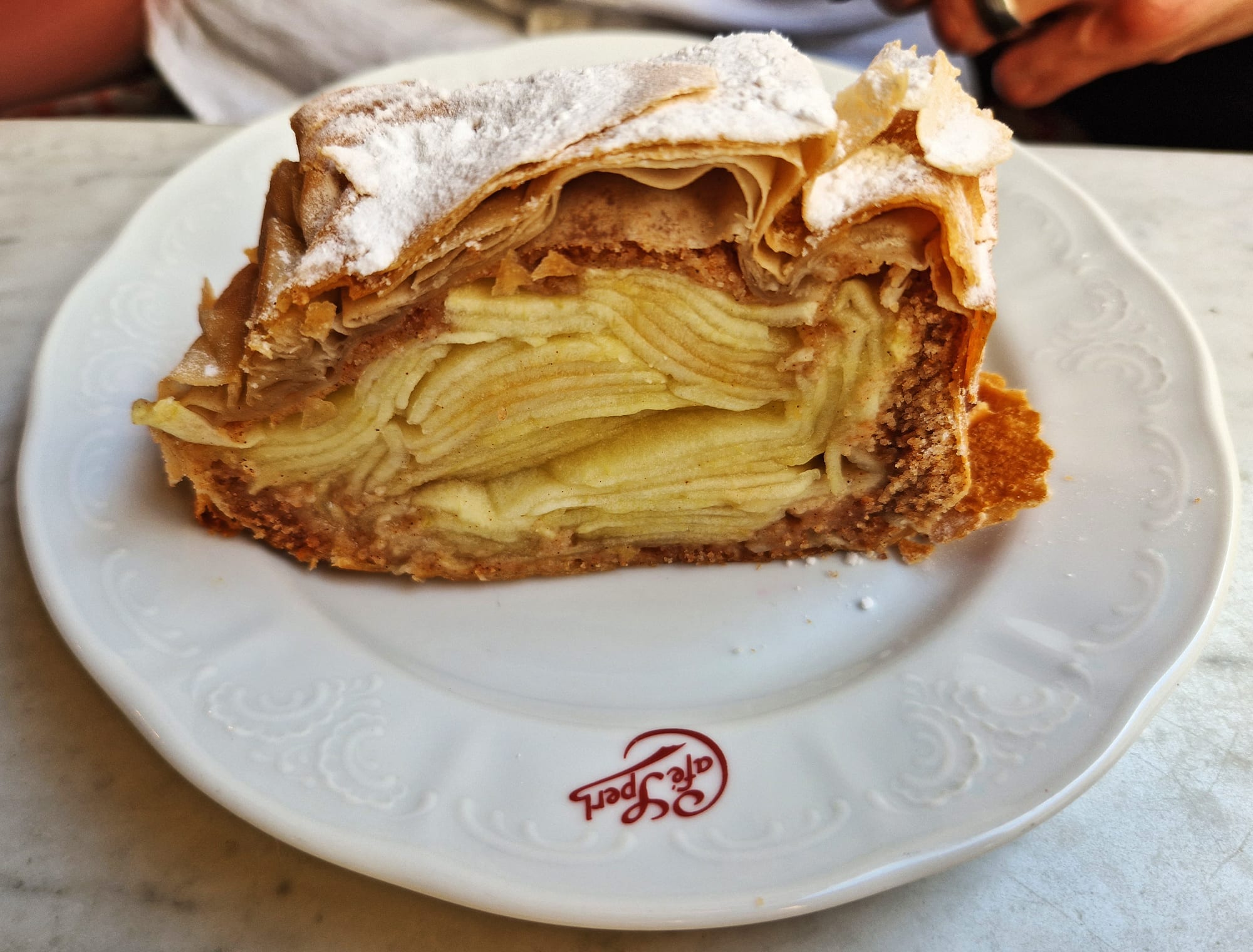A cross-section slice of strudel showing thin pastry layers wrapped around pale sliced apple, dusted with icing sugar