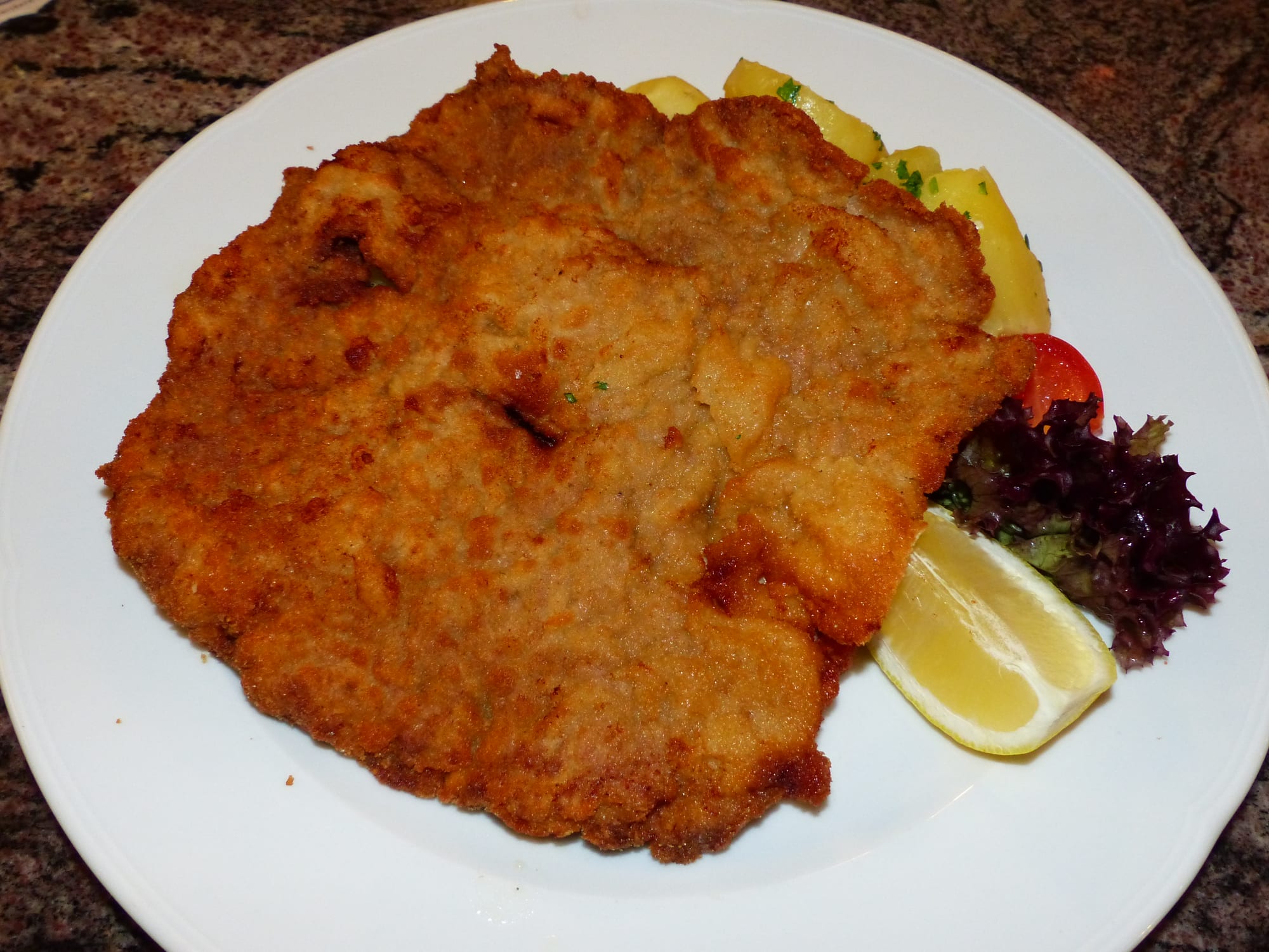 A large golden breaded veal cutlet filling most of a plate, with buttered parsley potatoes, a lemon wedge and salad on the side