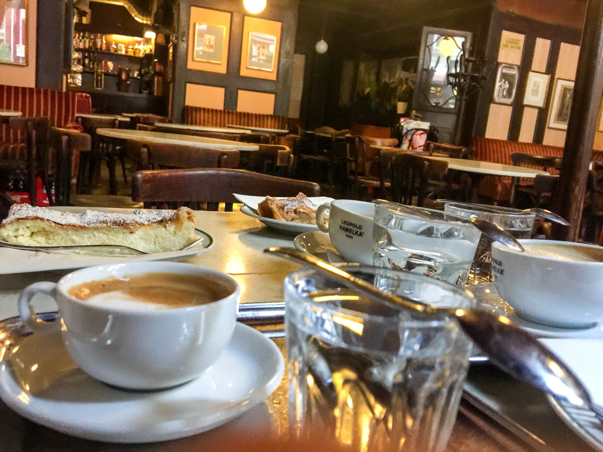 A dimly lit, old-fashioned coffeehouse interior with red banquette seating, framed pictures on the walls, and coffee and cake on a marble table in the foreground