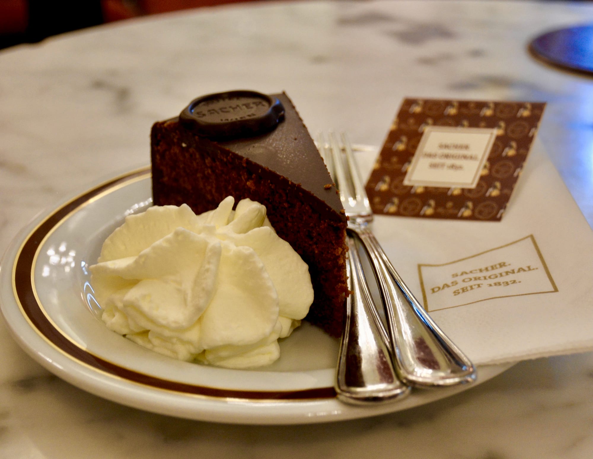 A wedge of dark chocolate cake with a round chocolate seal on top, served with a swirl of whipped cream