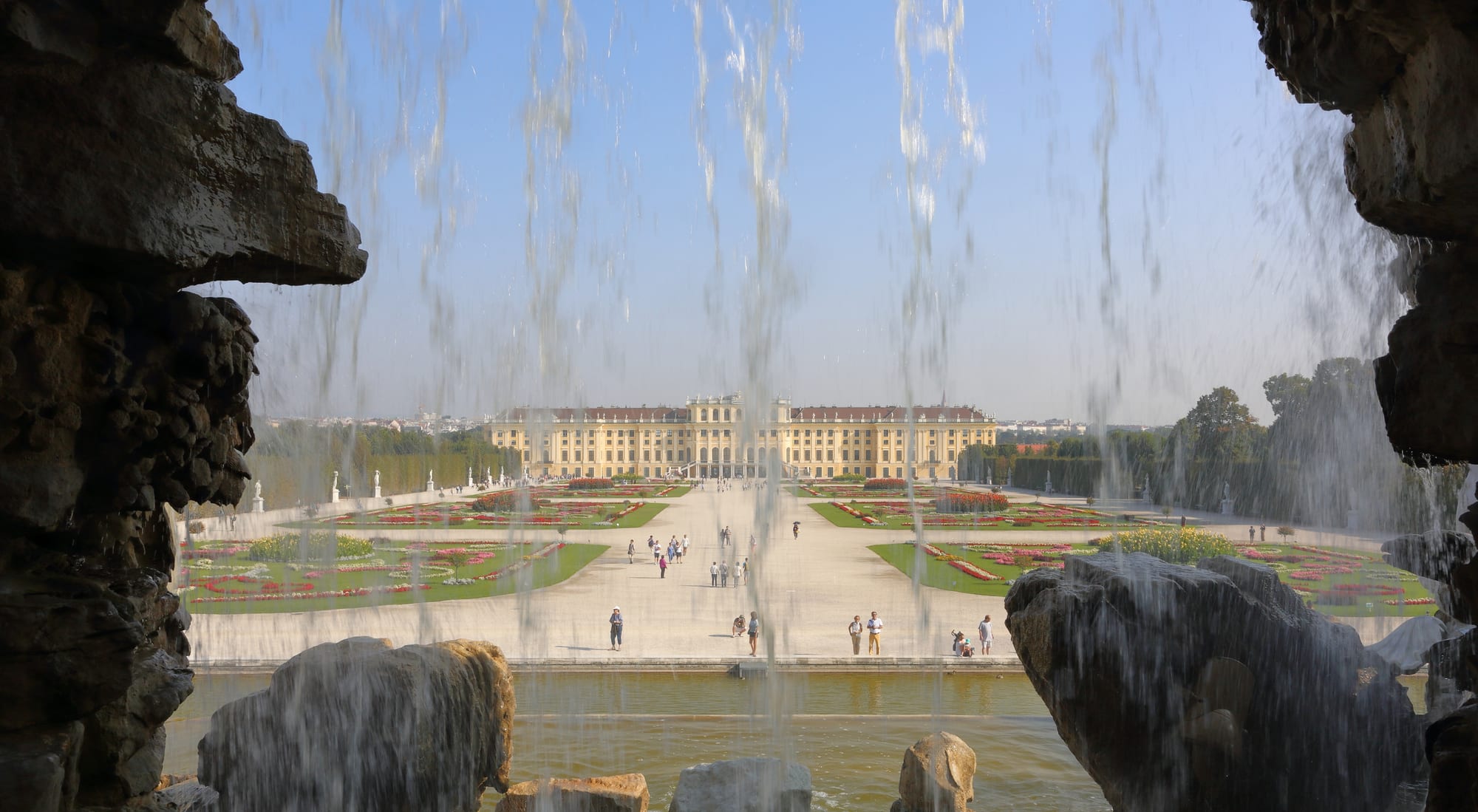 A long yellow baroque palace seen across formal gardens from a fountain terrace