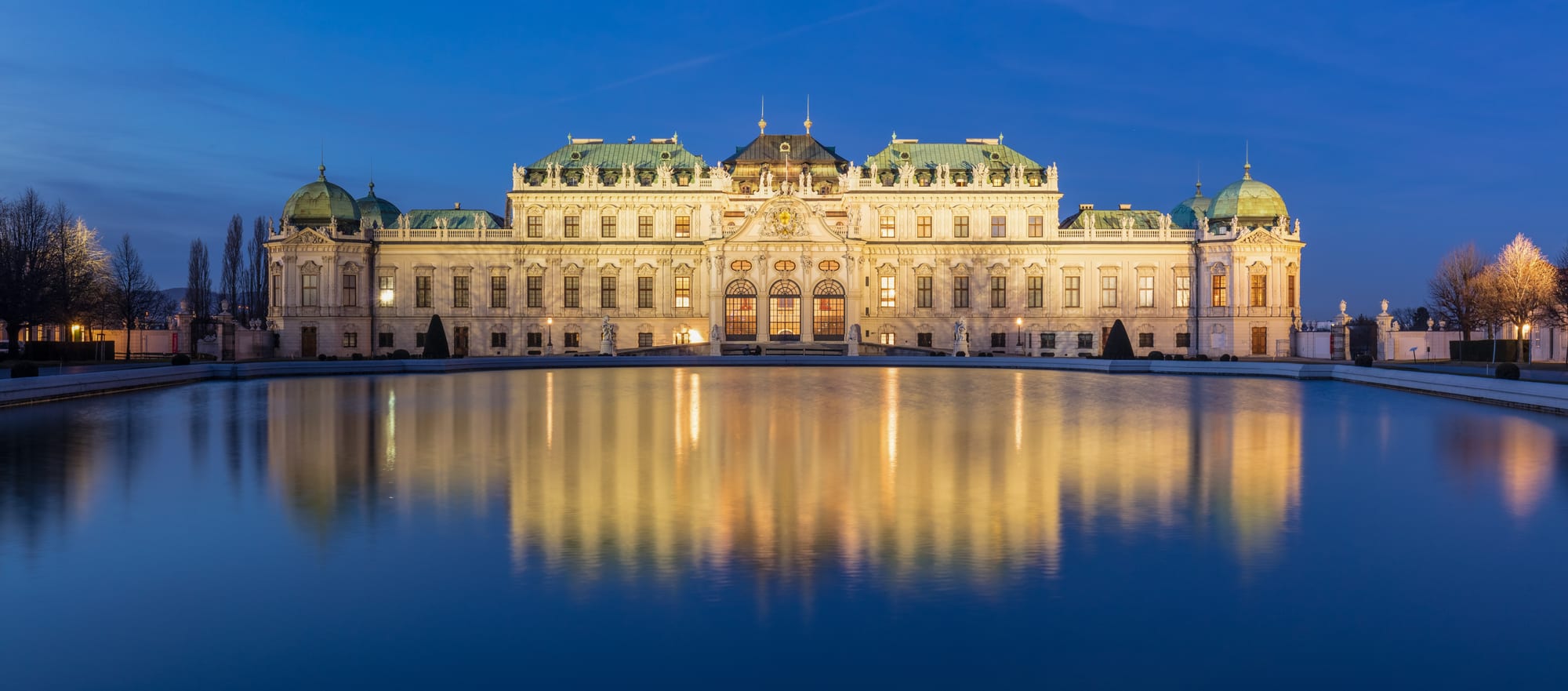 A baroque palace of white and gold behind a long ornamental pool and clipped gardens
