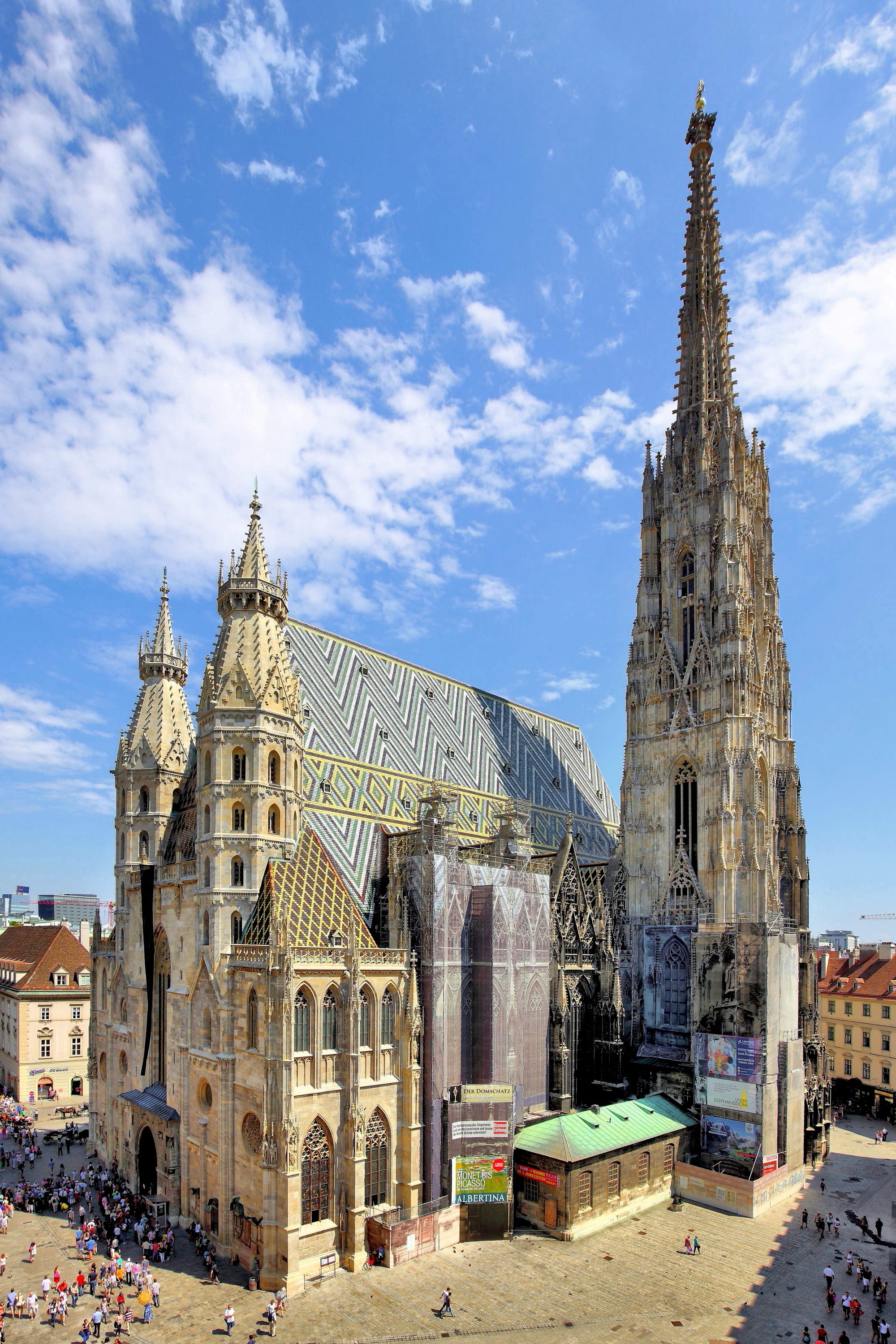 A vast Gothic cathedral with a steep patterned tile roof and a soaring stone spire, hemmed in by city buildings
