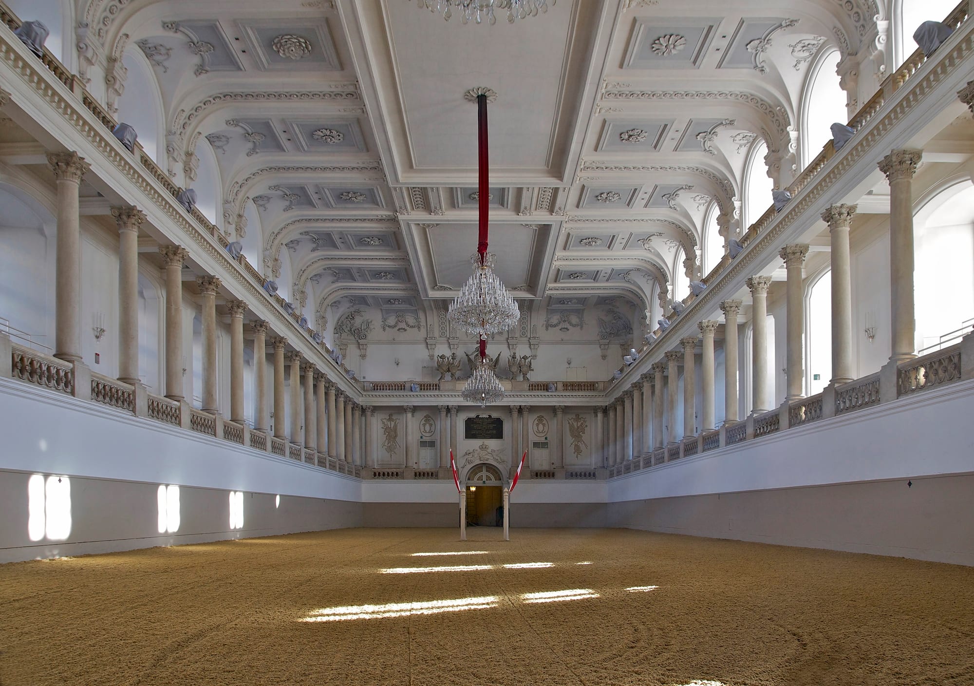 A long, empty white riding hall with a sand floor, columned galleries on both sides, an ornate coffered ceiling and crystal chandeliers