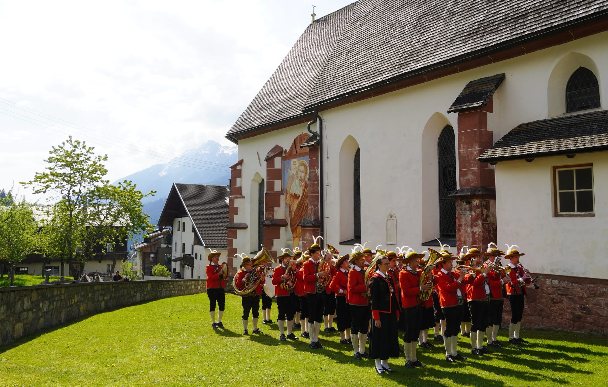A brass band in red jackets, black breeches and feathered hats lines up with its instruments on the grass beside a white village church, with snow-capped mountains behind