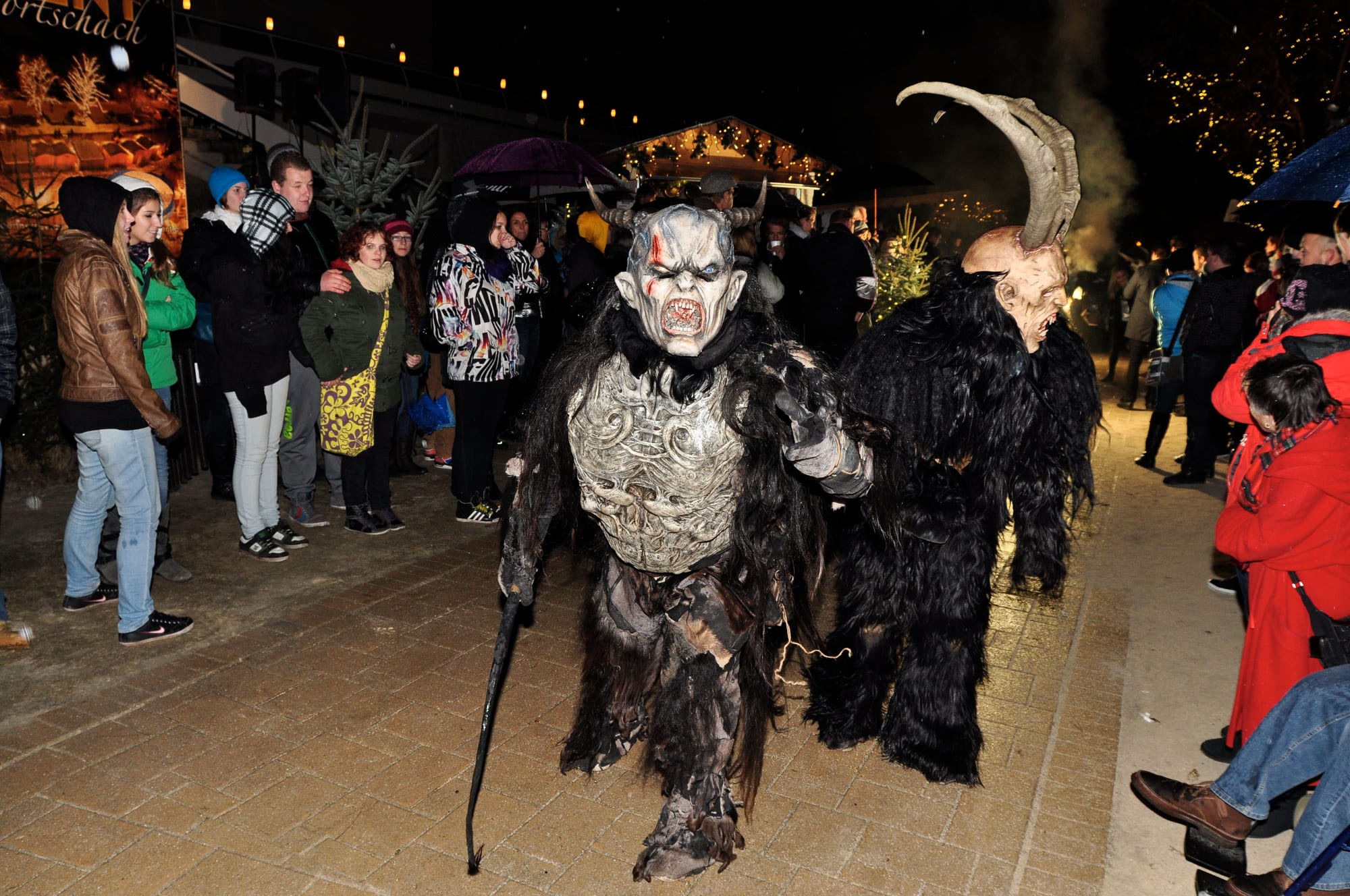 Two Krampus figures in shaggy black fur, carved horned masks and armour-like chest pieces walk along a paved promenade at night, watched by a crowd in winter coats