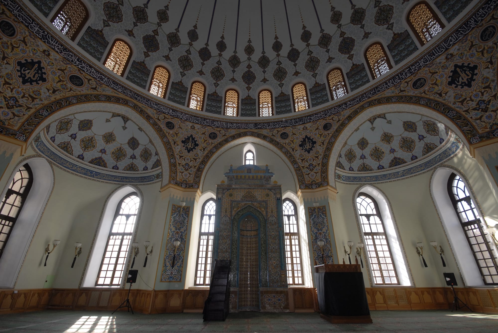 The interior of a mosque with a large dome painted in blue, gold and cream patterns, arched windows letting in sunlight, a tiled mihrab niche and a wooden pulpit, and an empty carpeted floor