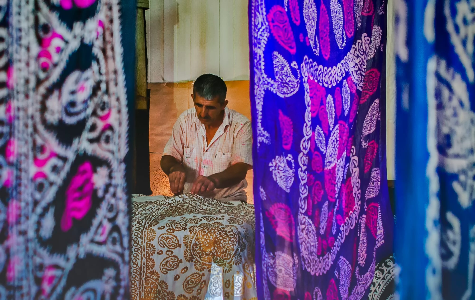 A craftsman at a table presses patterns onto a length of pale silk, framed between hanging silk scarves printed with bold purple, blue and pink paisley designs