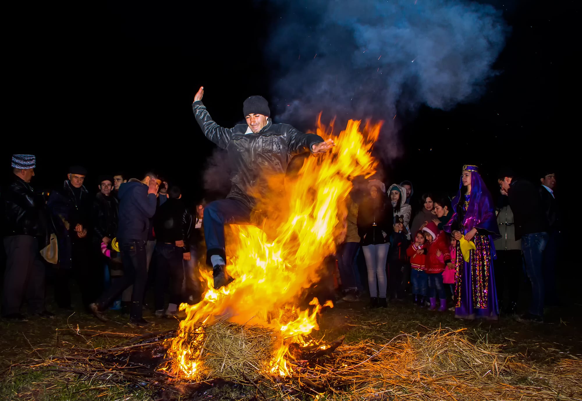 A man in a dark jacket and woollen hat leaps over a blazing straw bonfire at night, watched by a crowd that includes a young woman in a sequinned purple costume