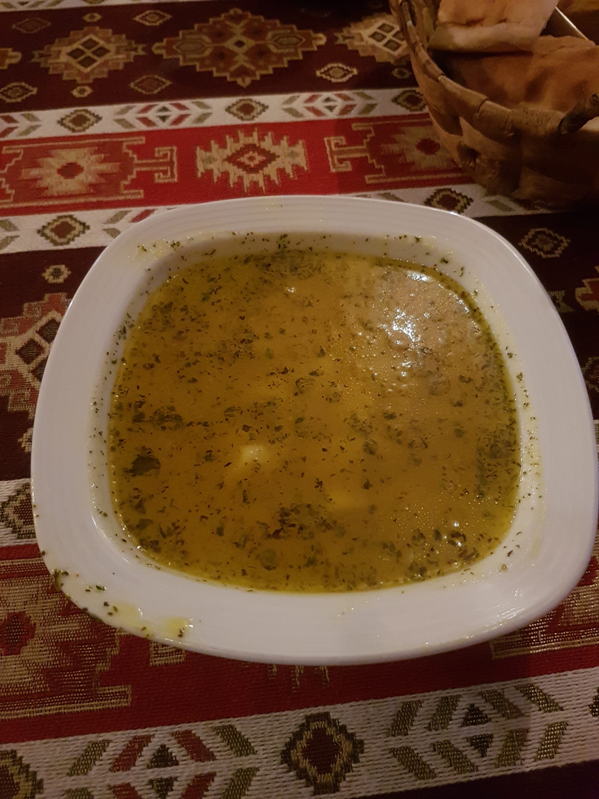 A square white bowl of clear, herb-flecked broth with small dumplings, next to a basket of bread