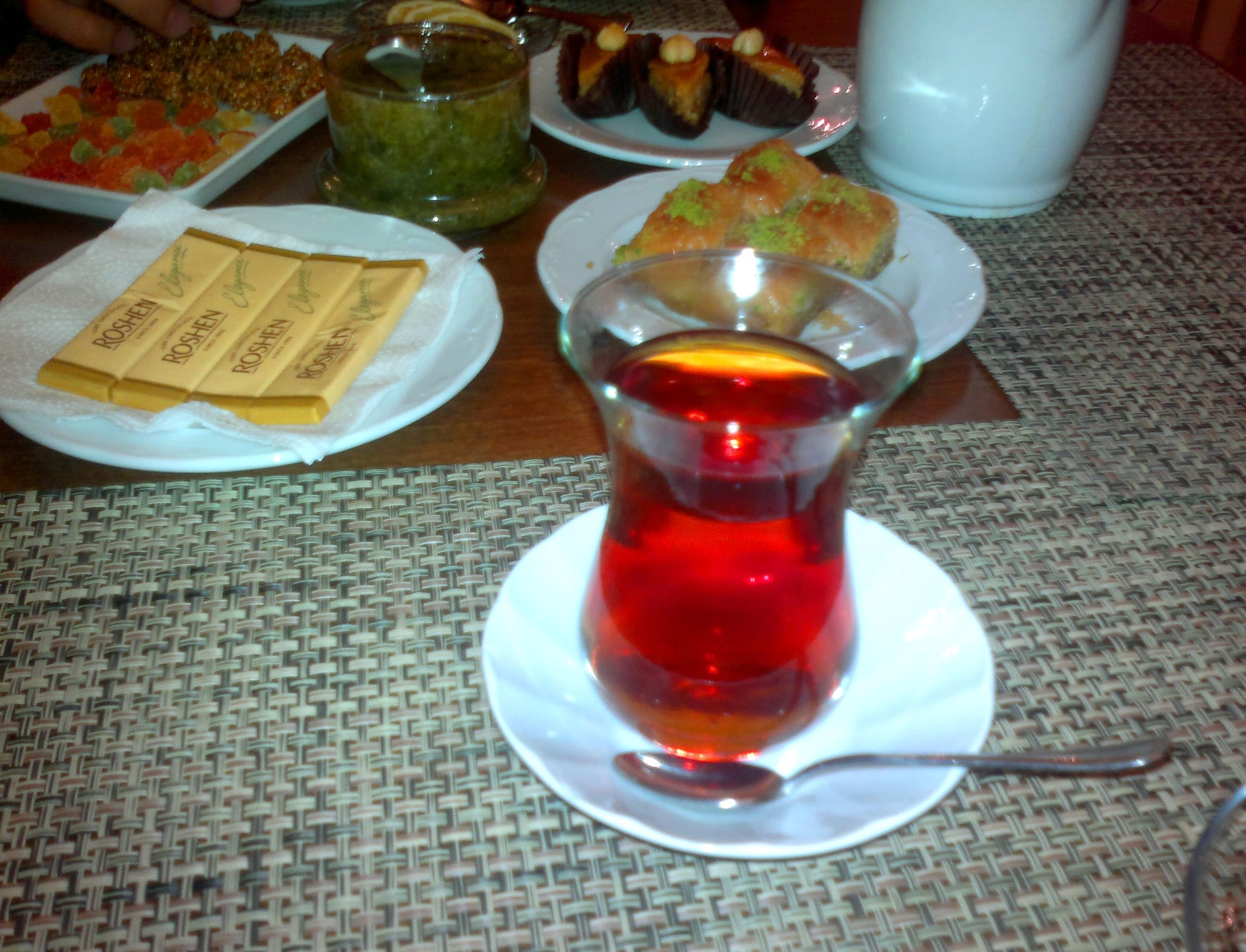 A pear-shaped glass of dark red tea on a saucer, with plates of pastries, jam, dried fruit and chocolate on the table behind