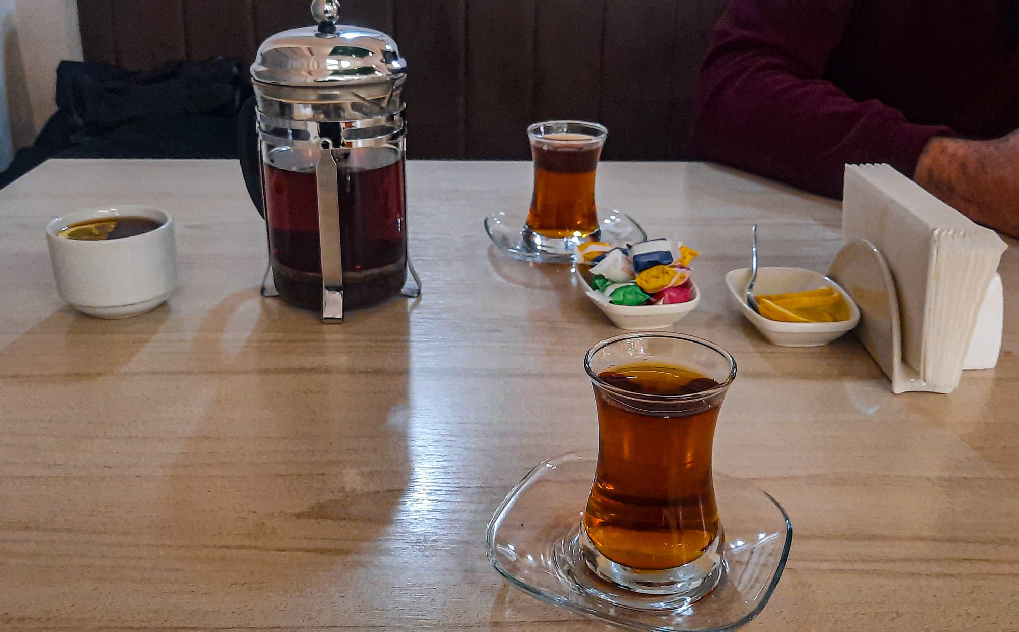 Two pear-shaped glasses of black tea on saucers next to a French press of tea on a café table, with a seated customer in the background