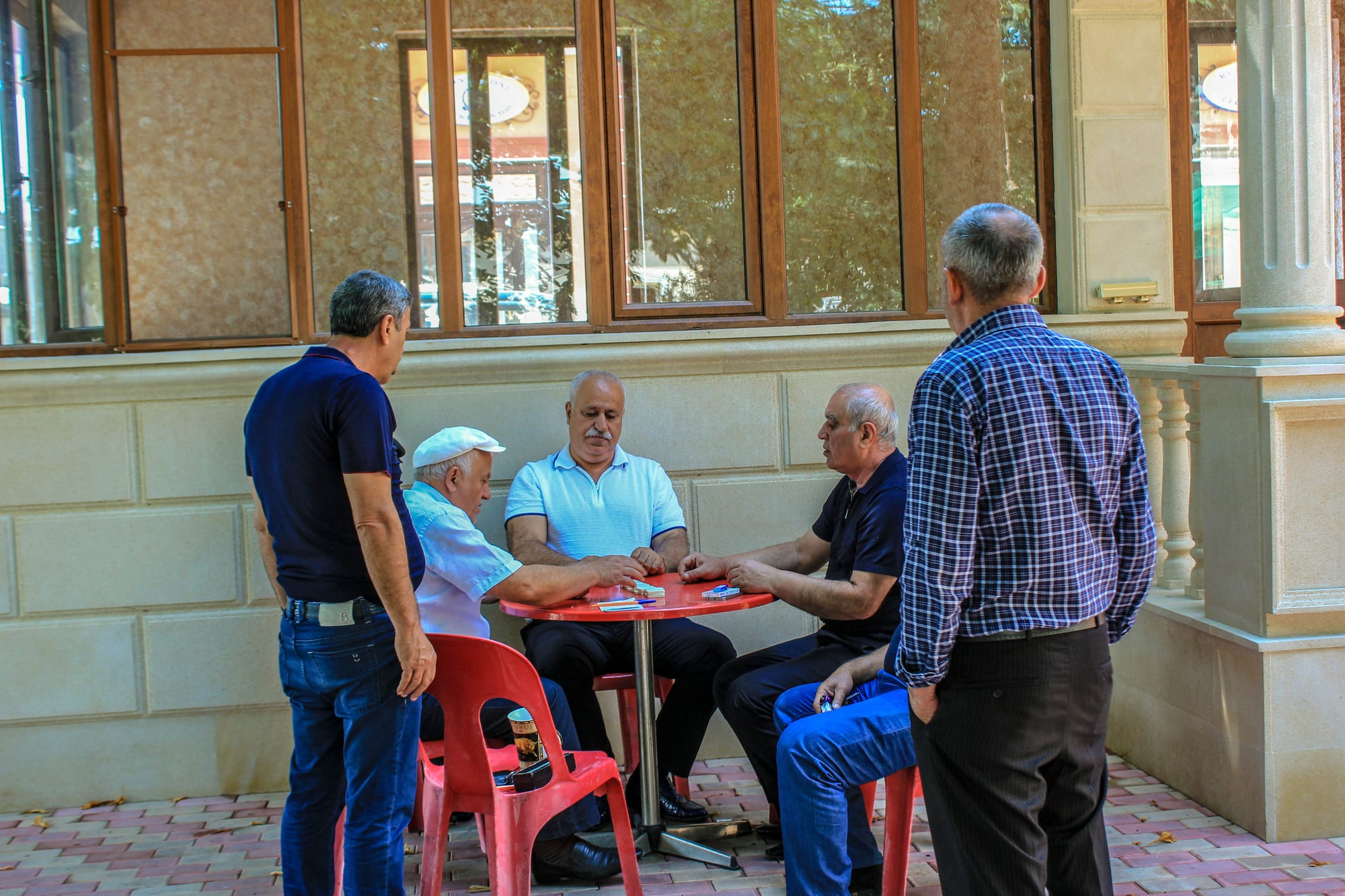 Five men gathered around a small red outdoor table at a teahouse, two of them seated and playing a table game while the others stand and talk
