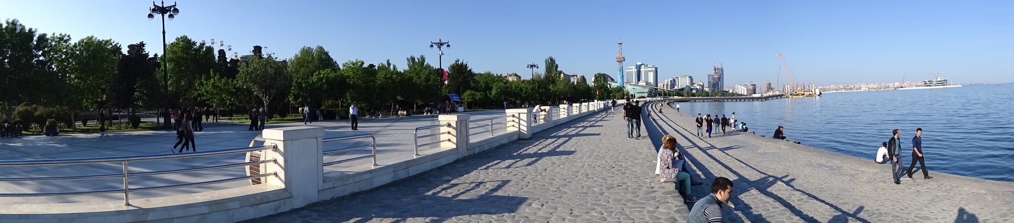 A wide paved seaside promenade with people walking and sitting along a railing beside the Caspian Sea, city towers in the distance