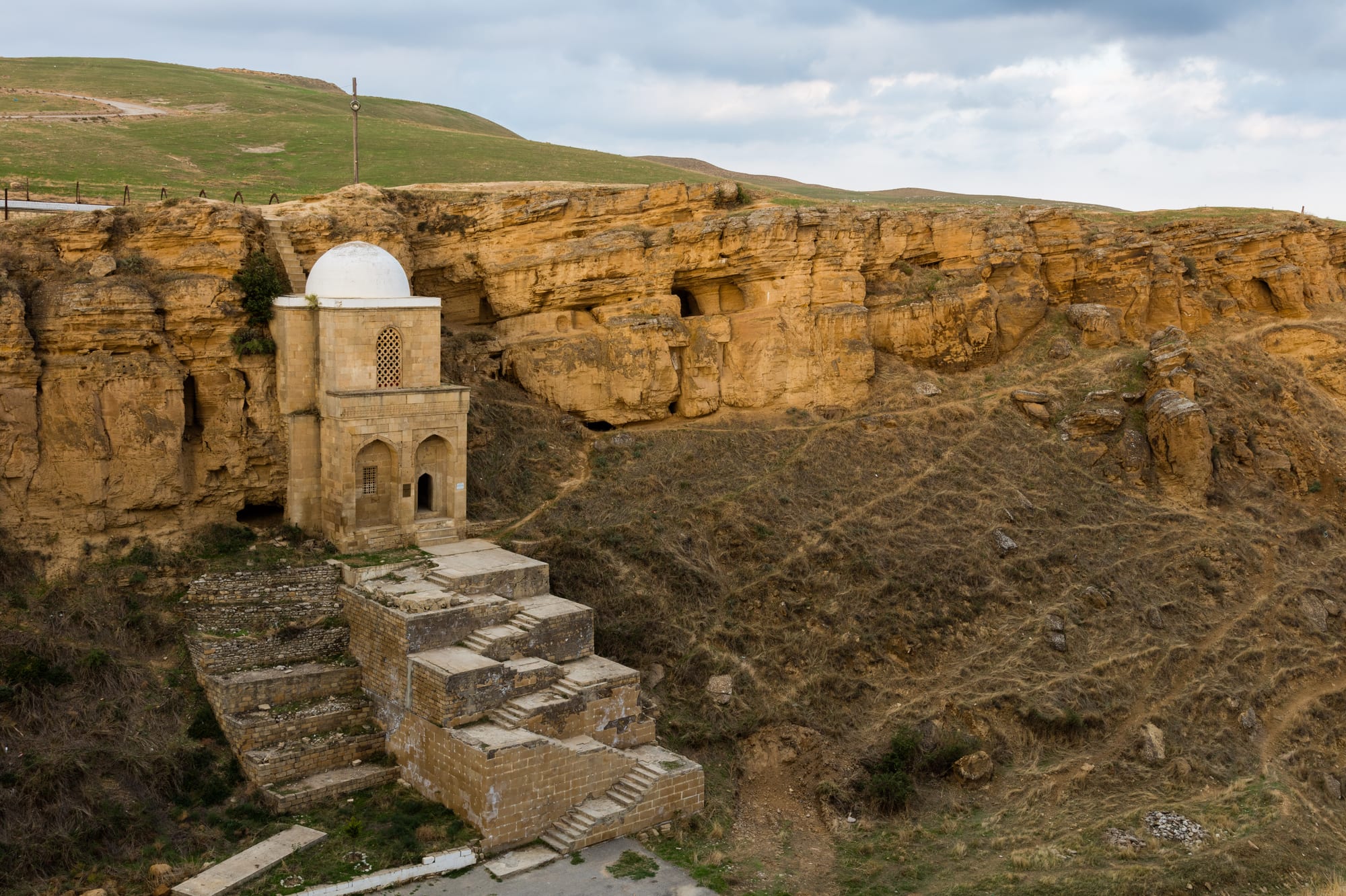 A small two-storey stone mausoleum with a white dome built against a golden sandstone cliff, with stone stairs climbing to it from the valley floor
